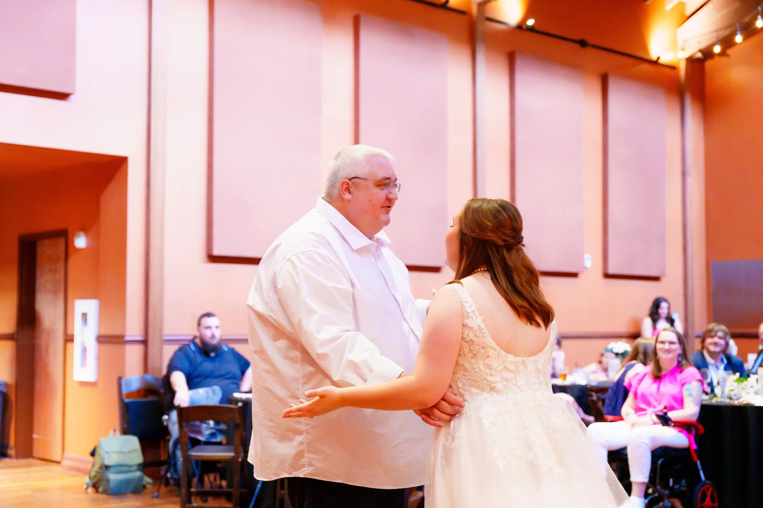 A couple dancing together at a wedding reception, with guests seated and watching in the background. The man has silver hair and glasses, and the woman has brown hair styled in loose waves. The venue has warm lighting and pinkish walls.