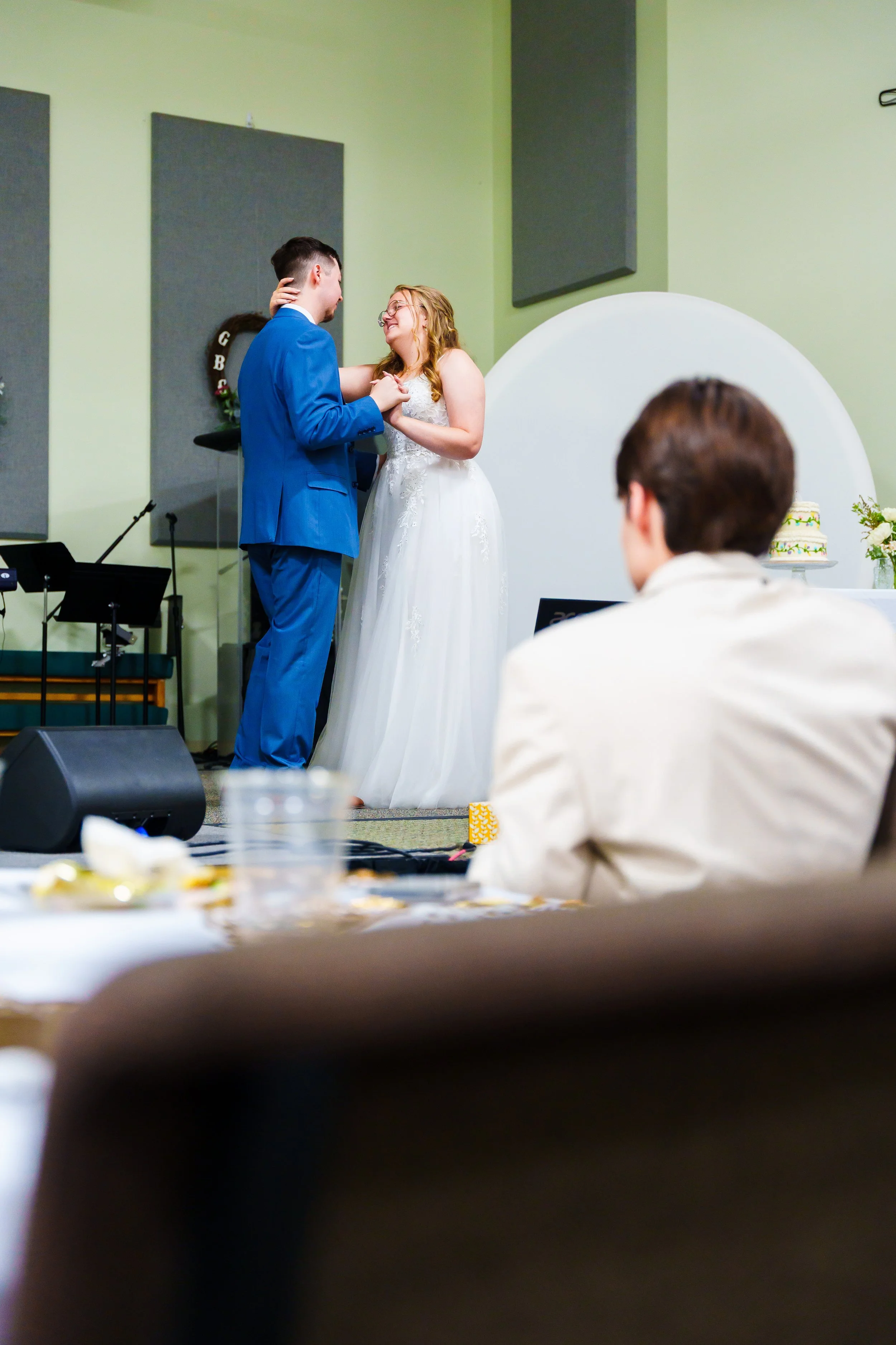 A wedding ceremony with a bride and groom dancing together in a room with green walls, while a guest watches from the foreground.
