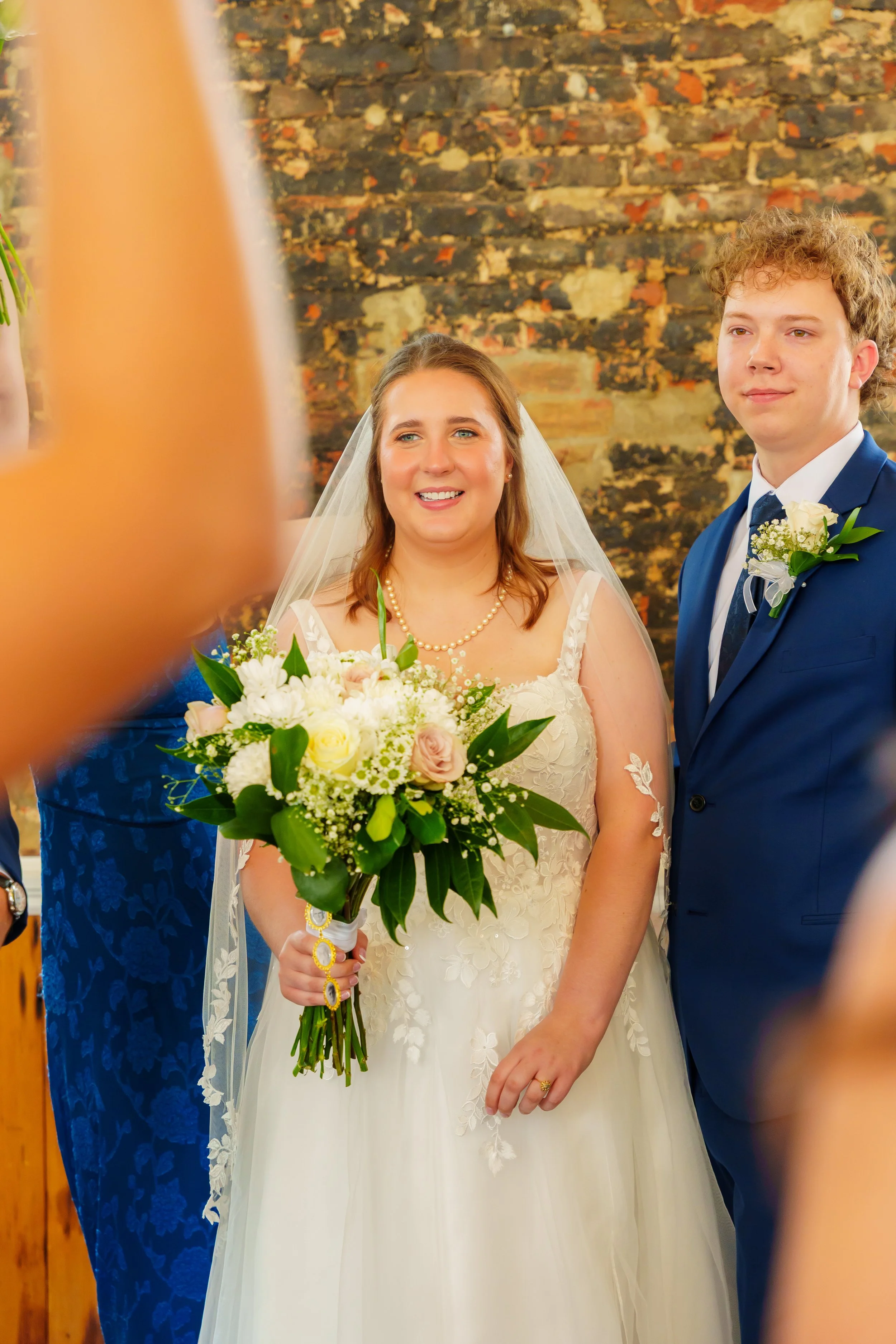 A bride in a white wedding gown holding a bouquet of white and pink flowers, standing next to a groom in a blue suit with a boutonniere, during a wedding ceremony.