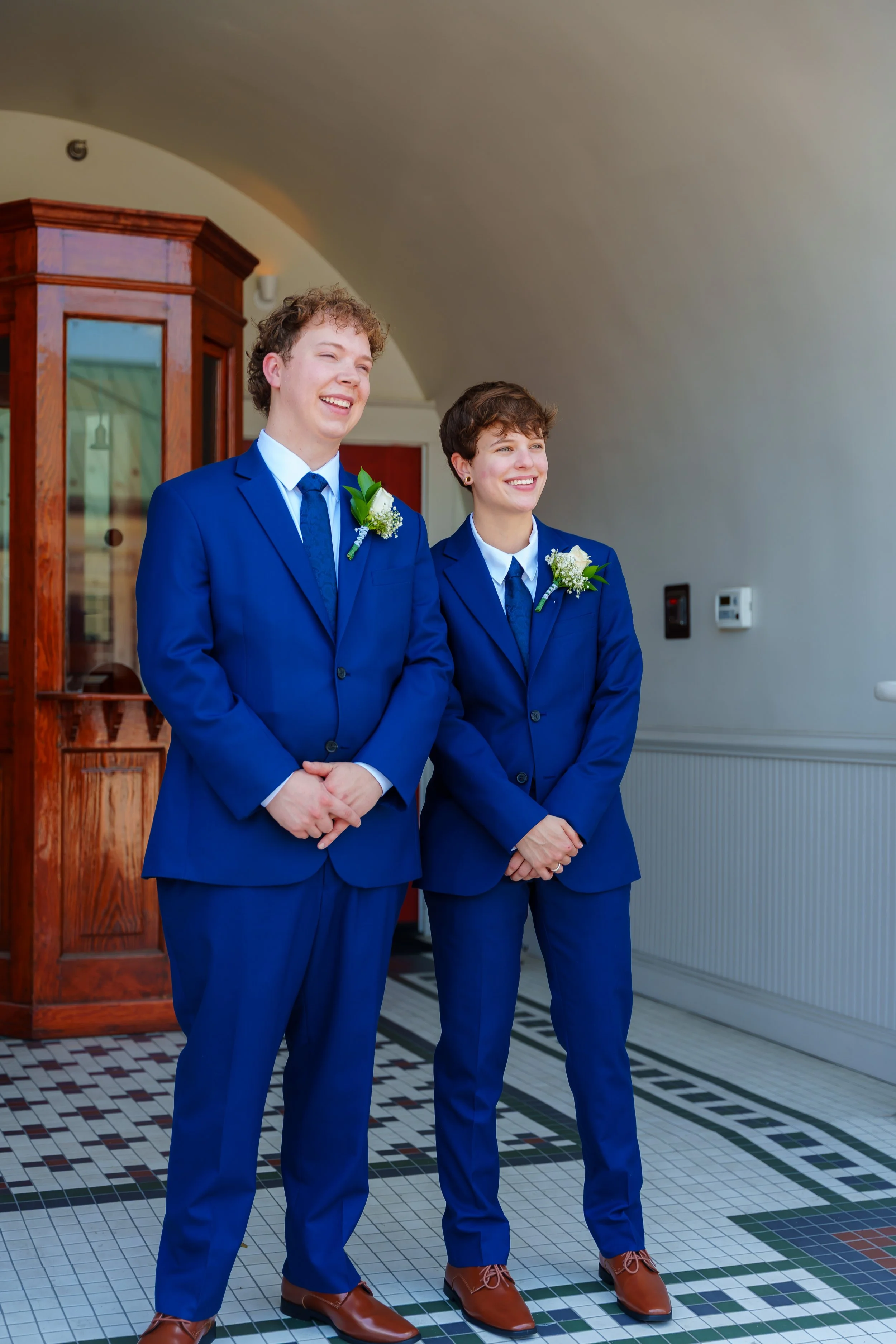 Two people wearing matching blue suits with white shirts and blue ties, standing indoors, each with a boutonniere, smiling. A wooden display cabinet is in the background, and the floor has a colorful tile pattern.