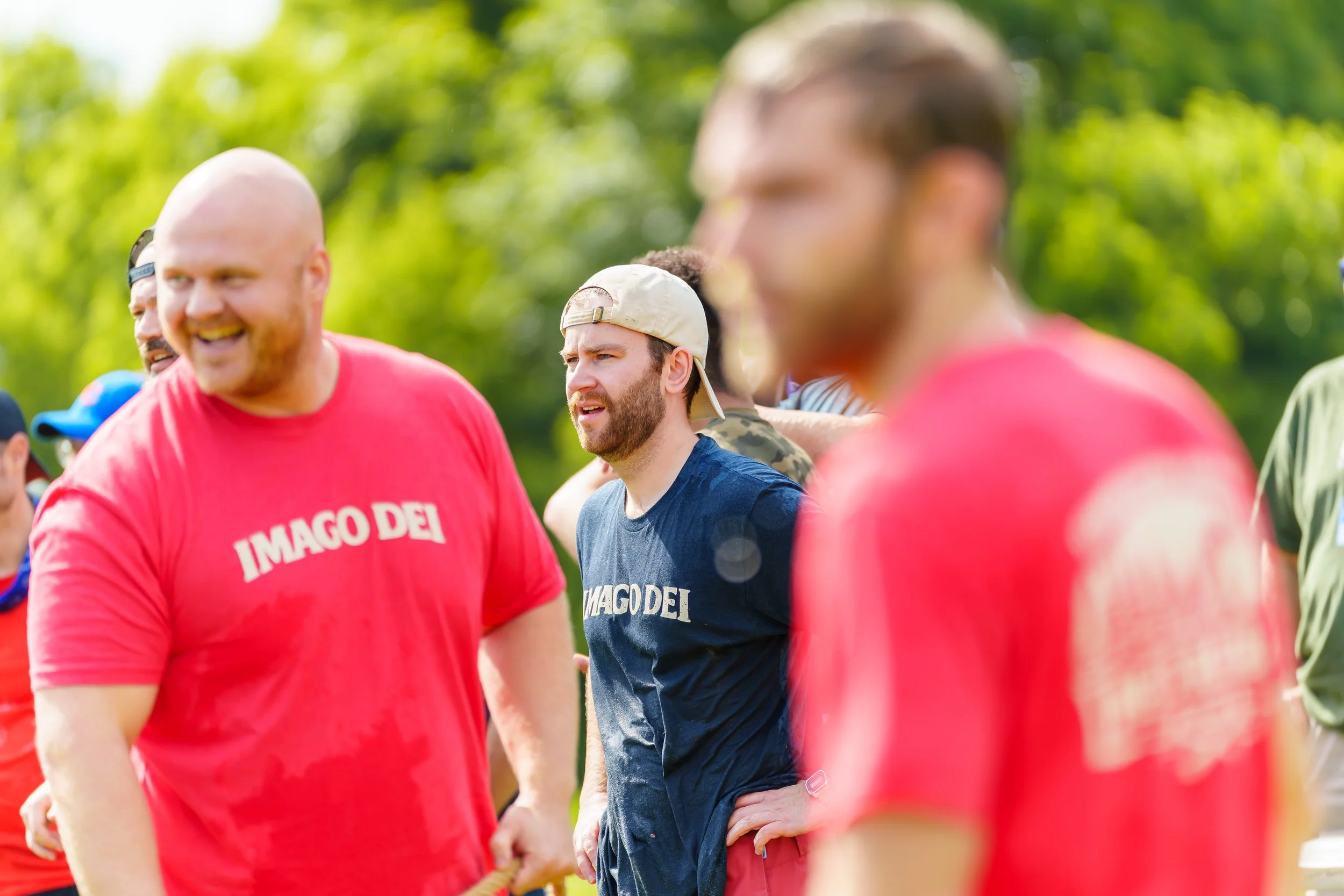 A group of diverse young men outdoors, smiling and engaging in a team activity, some wearing casual clothing with one in a red shirt that has white text, and others in blue or camouflage attire, with green trees in the background.