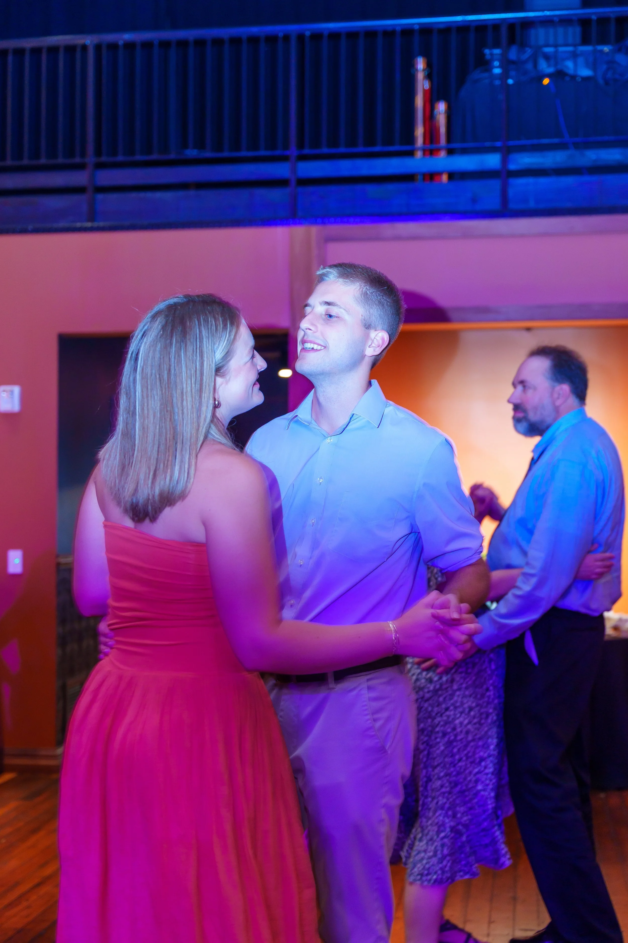 Couple dancing together at a social event, with a man smiling at a woman wearing a pink dress. In the background, another couple is seen dancing and talking.