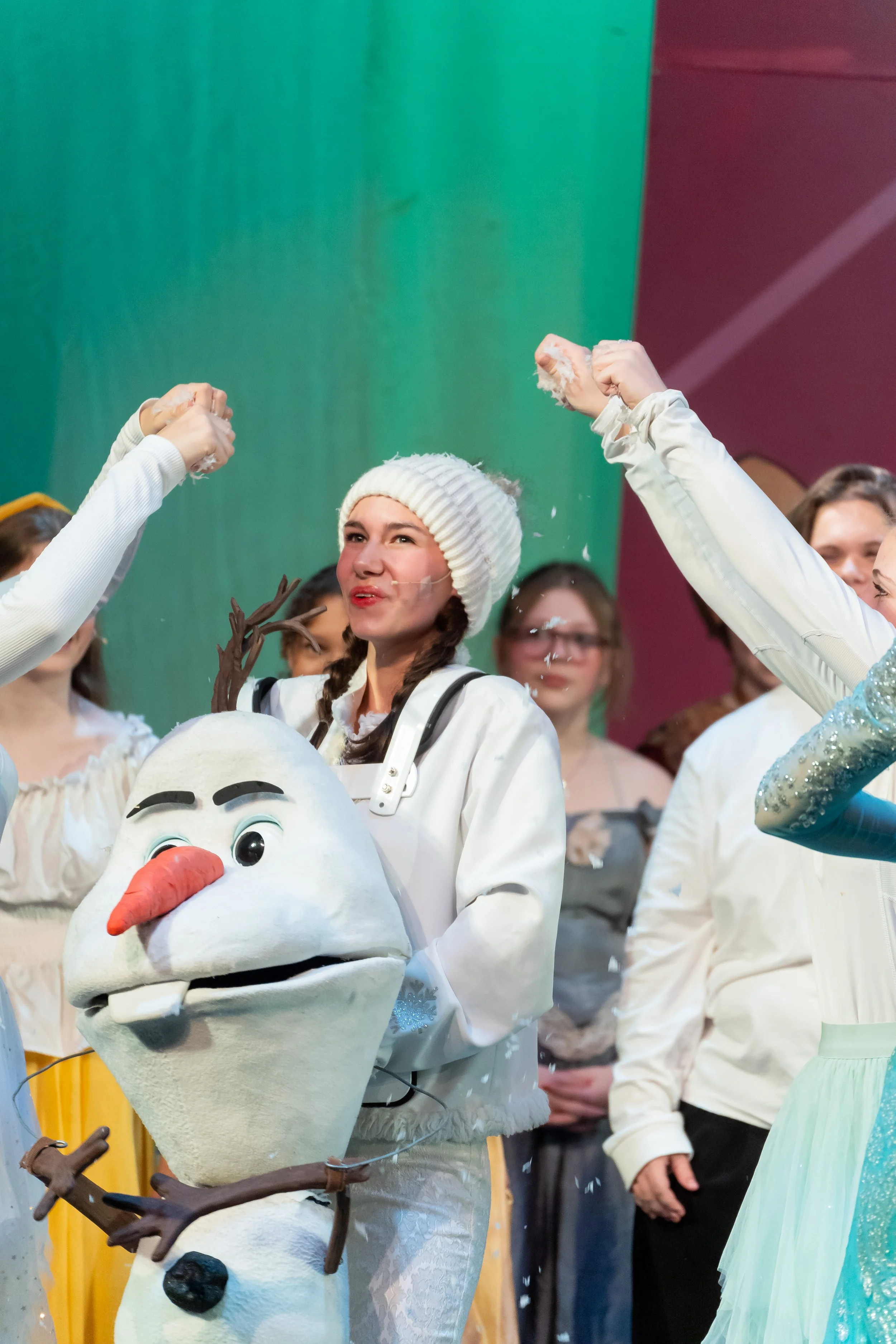 A woman dressed as Olaf from Frozen is holding a large Olaf puppet at a festive event. She is wearing a white knit hat and a white costume, with other people in the background.