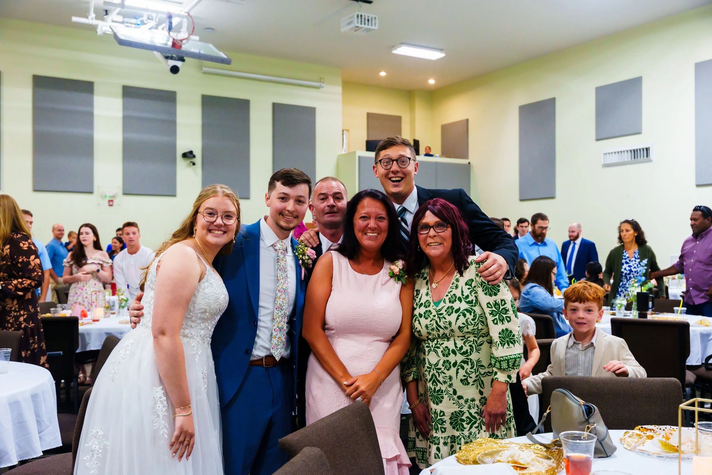 Group of people at a celebration, with five individuals posing for a photo in the foreground, including a woman in a pink dress, a man in a blue suit, someone in a white wedding dress, a woman in a green floral dress, and a tall man in a black suit w