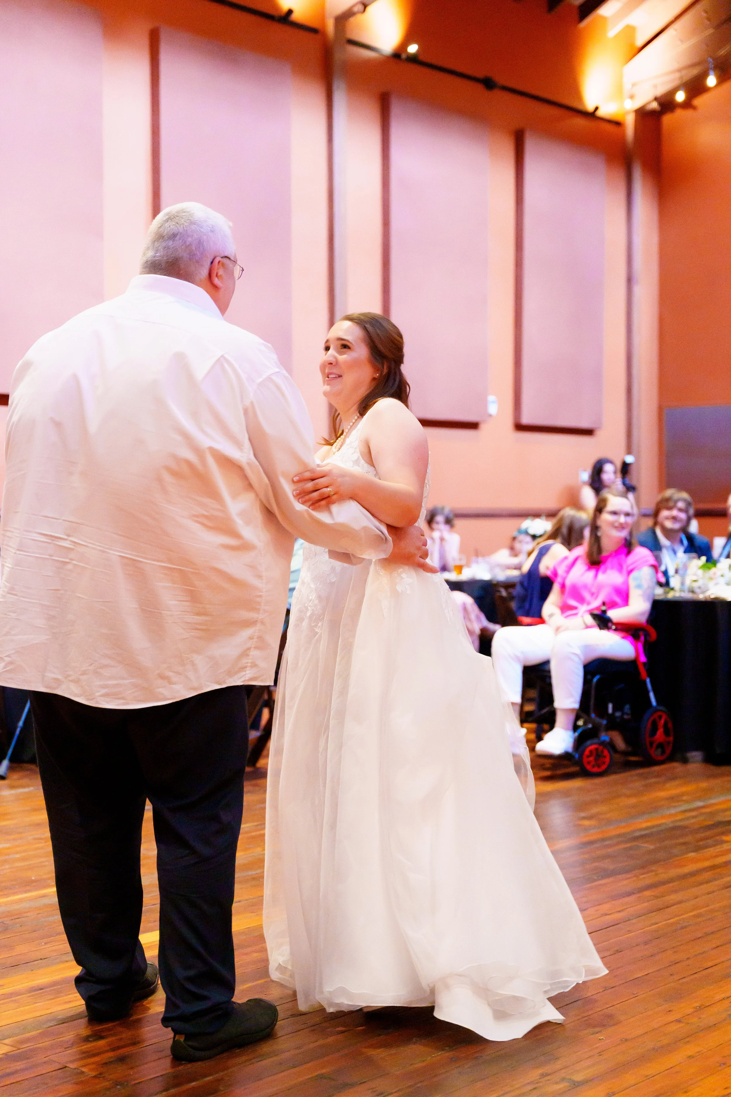 A bride and an older man dance together at a wedding reception in a decorated banquet hall with guests sitting and watching in the background.