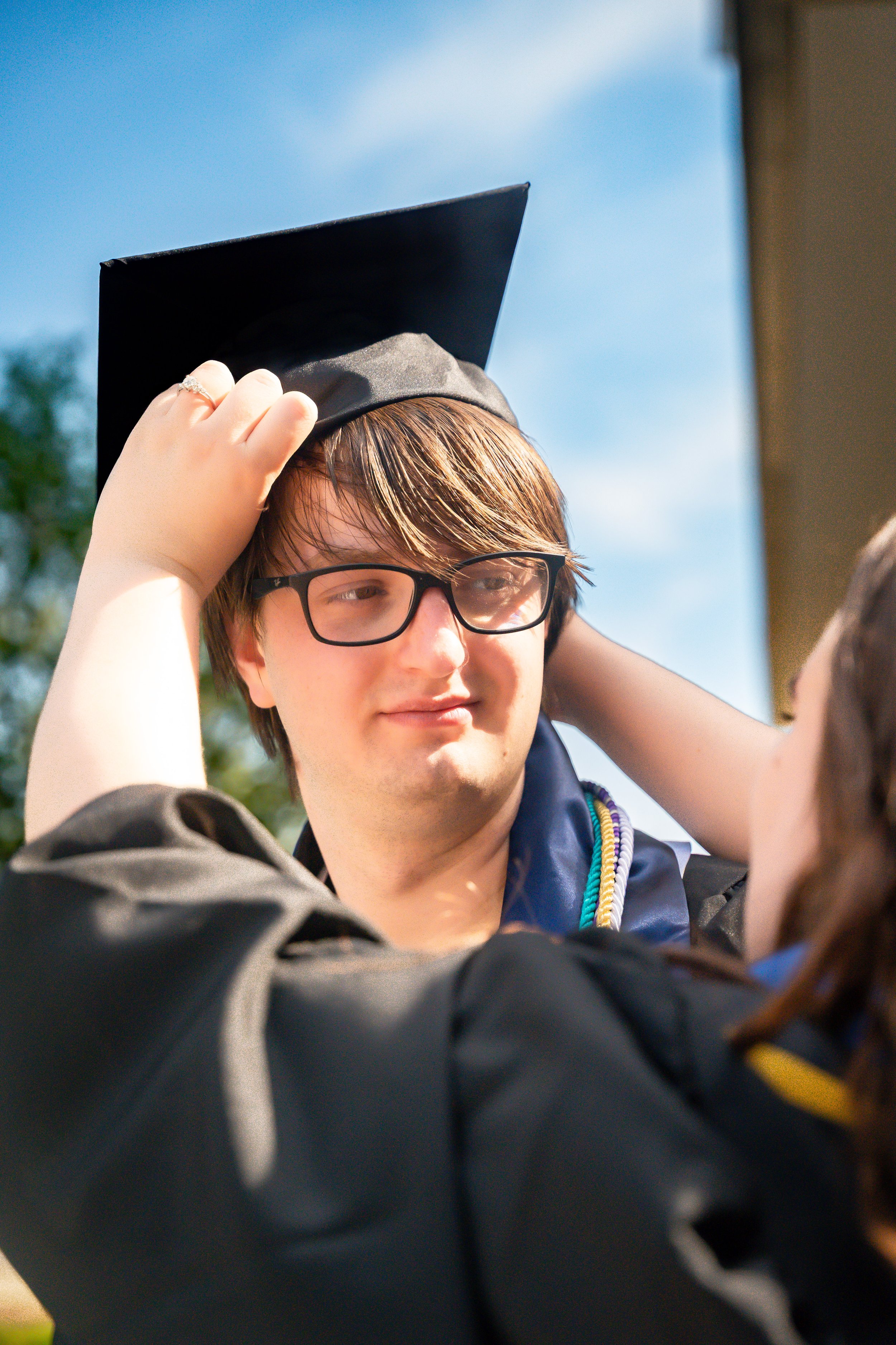 A young man with glasses and brown hair wearing a graduation cap and gown, adjusting his cap outdoors on a bright, sunny day.