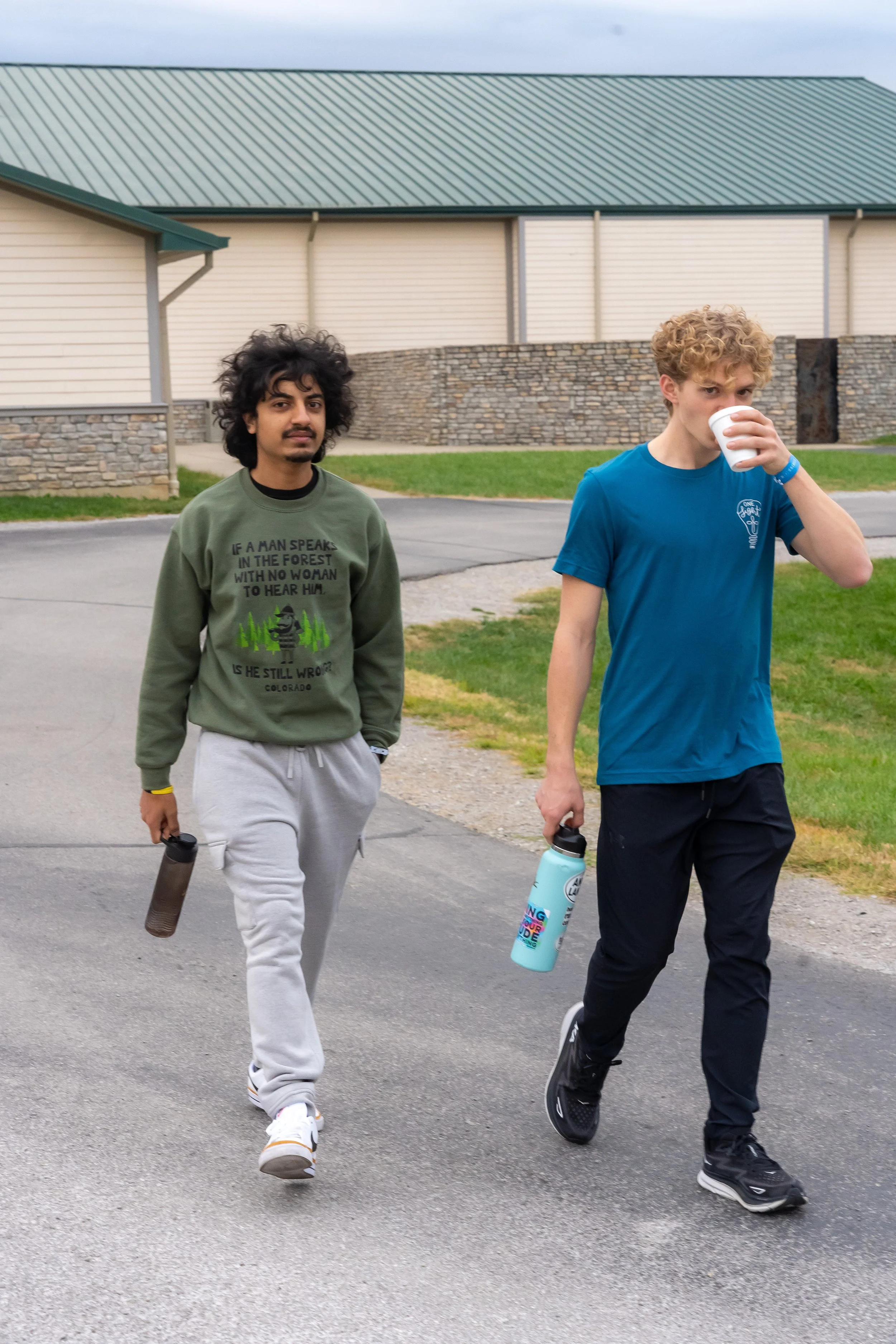 Two young men walking outdoors in casual clothing, each holding a water bottle. One has curly black hair and a green sweatshirt with printed text, the other has curly blonde hair and a blue t-shirt.