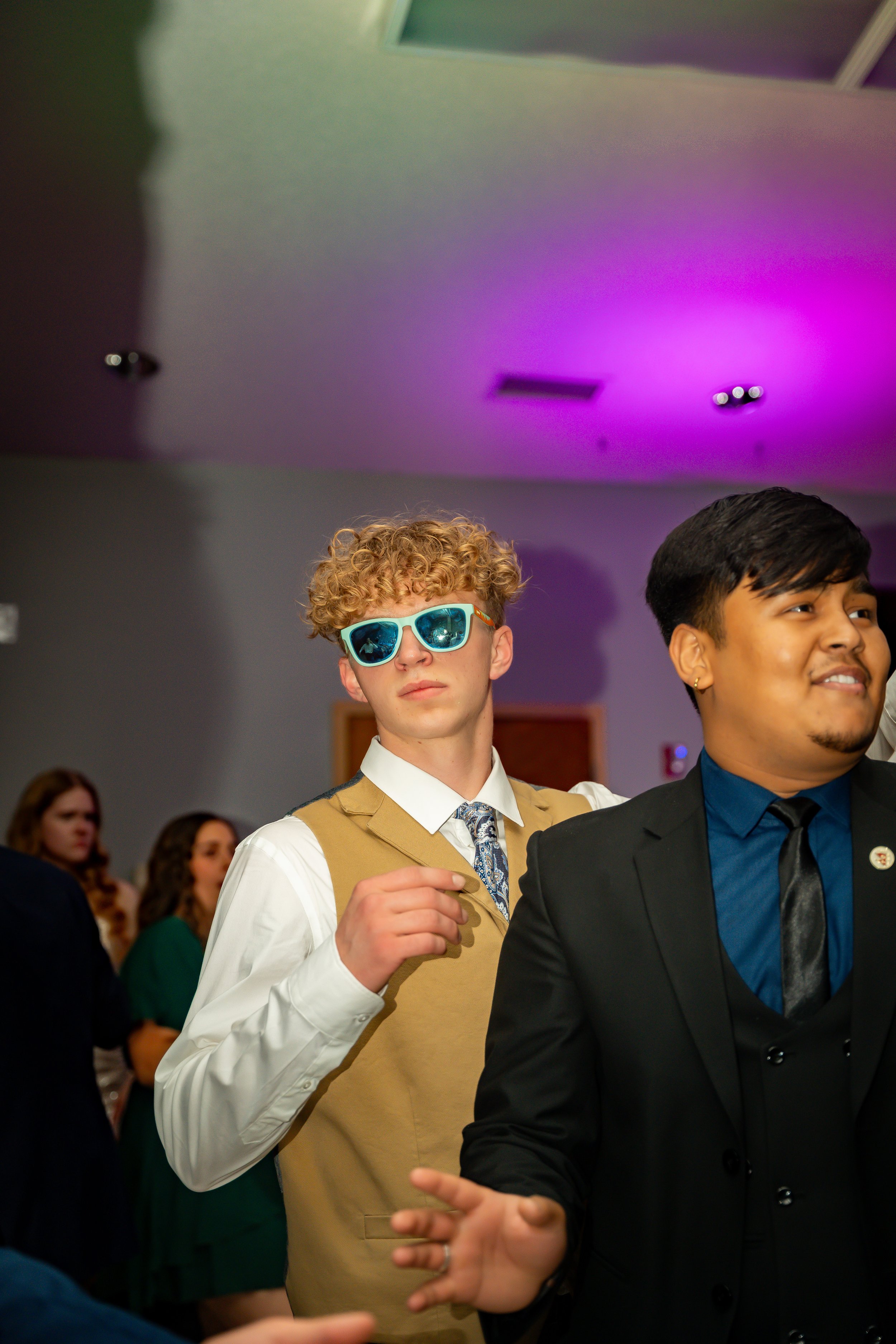 Two young men at a social gathering. One with curly blonde hair wearing blue sunglasses, white shirt, tan vest, and a patterned tie. The other with black hair, wearing a dark suit with a blue shirt and black tie. People in the background are blurred,