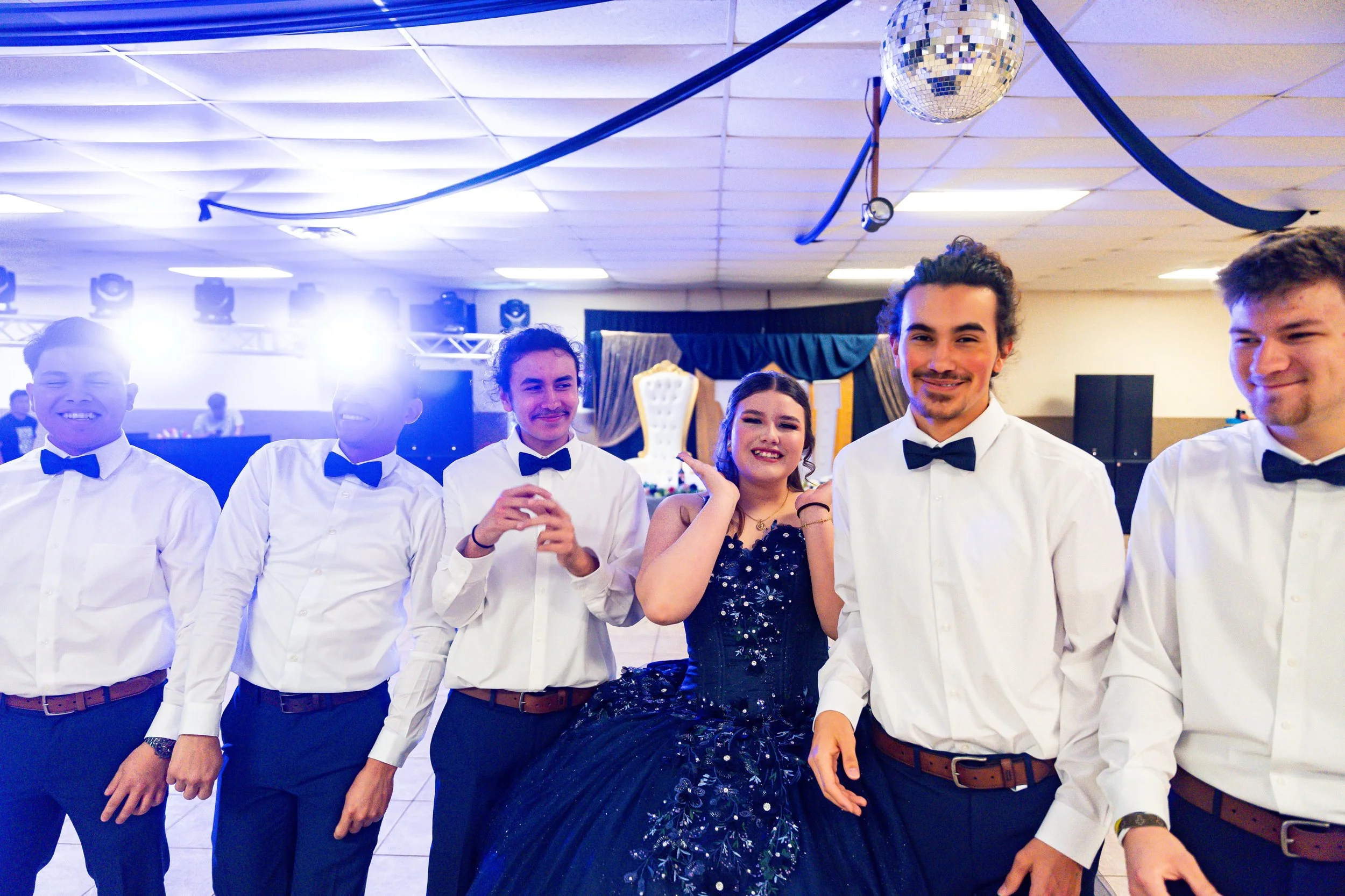A group of five young people dressed in formal attire, standing side by side at an indoor event with a ceiling decorated with streamers and a disco ball, smiling and posing for the photo.