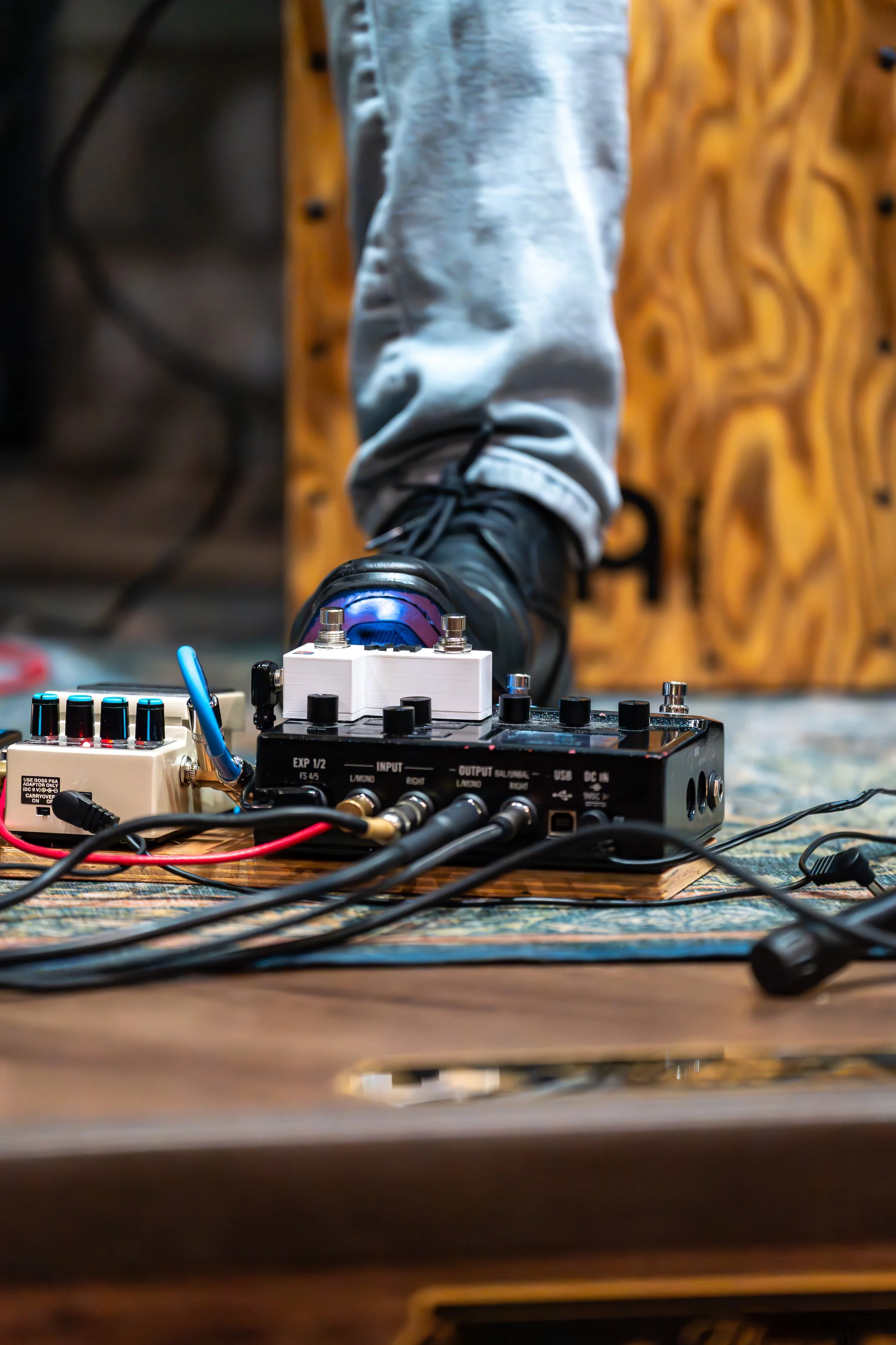 Close-up view of a foot wearing a black boot stepping on a guitar effects pedal with various knobs and cables scattered around, on a wooden floor.