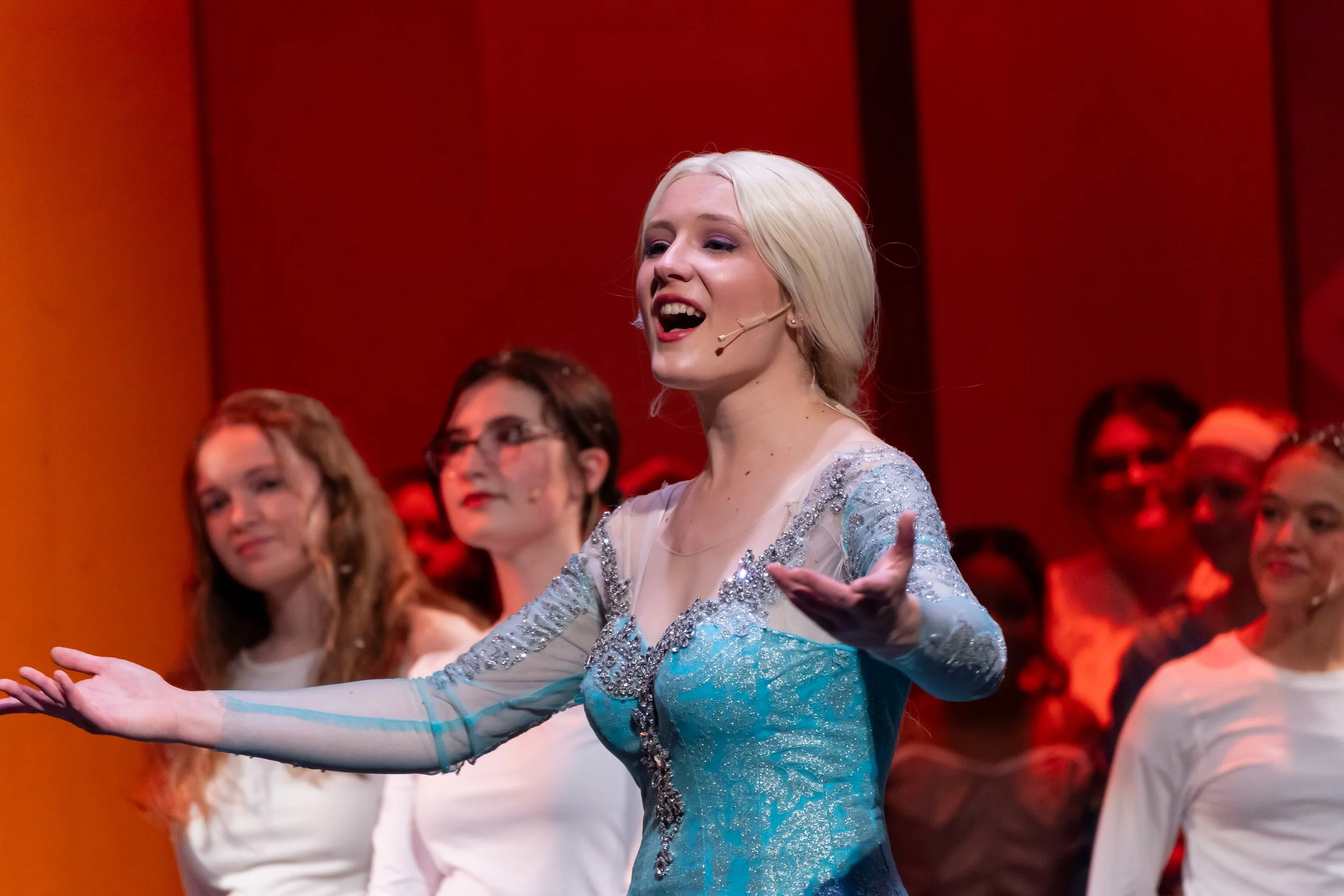 A woman with platinum blonde hair singing or performing on stage, wearing a sparkly blue dress with sheer sleeves, surrounded by others in the background.