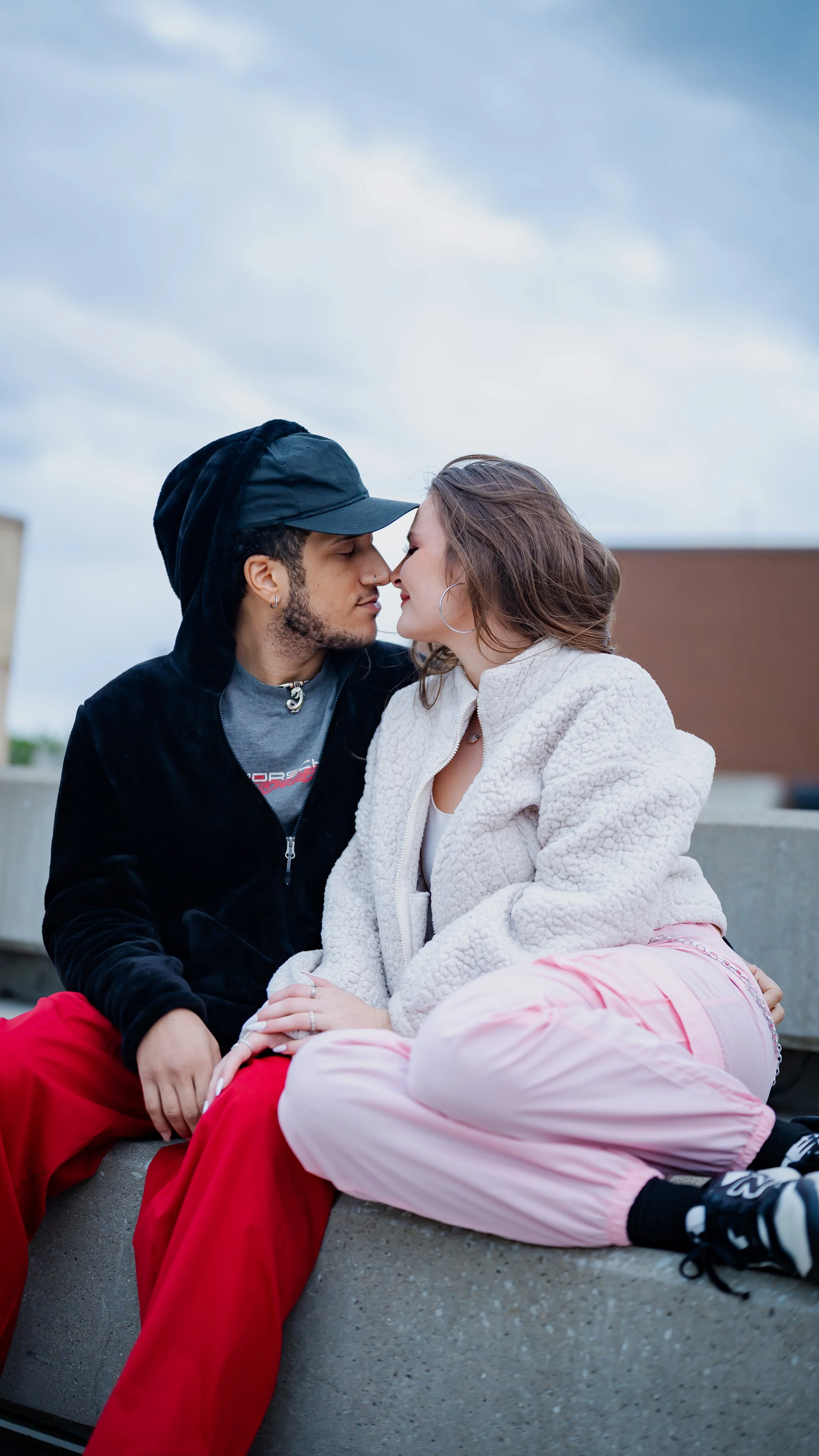 A couple sitting close together outdoors, touching foreheads and smiling. The man is wearing a black jacket, hoodie, and red pants; the woman is wearing a cream fleece jacket, pink pants, and sneakers.