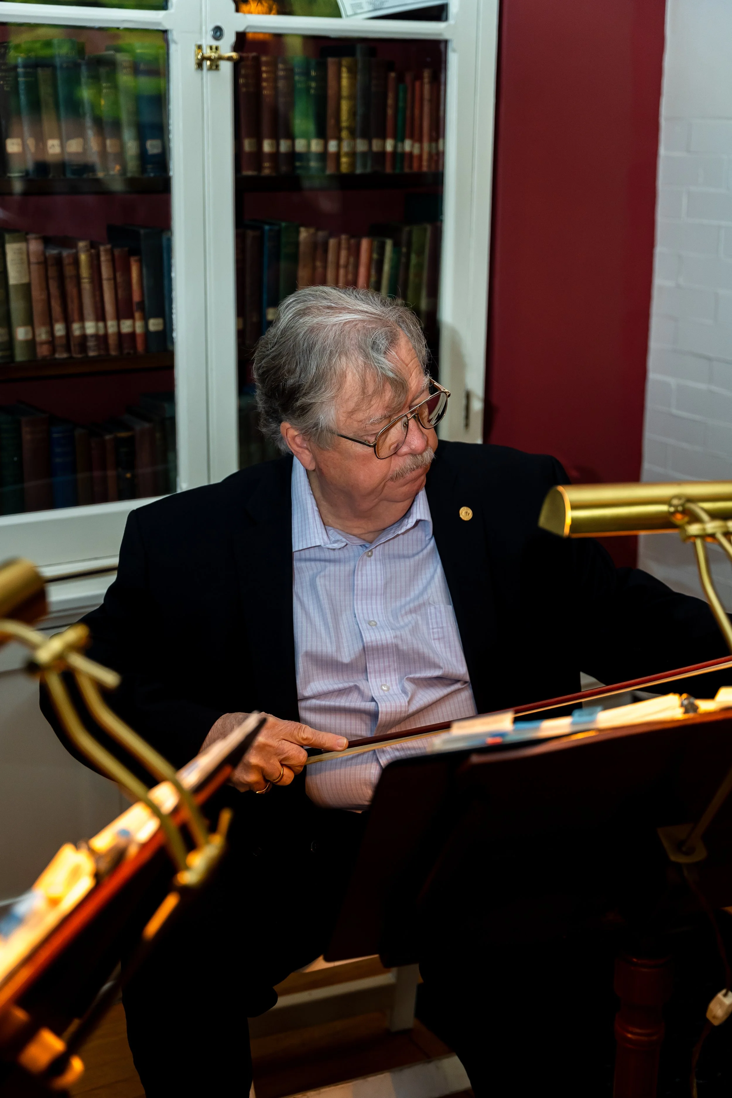 An elderly man with gray hair, glasses, in a black suit, sitting at a piano, in a room with a bookshelf filled with books behind him, and unique gold-colored piano lamps in the foreground.