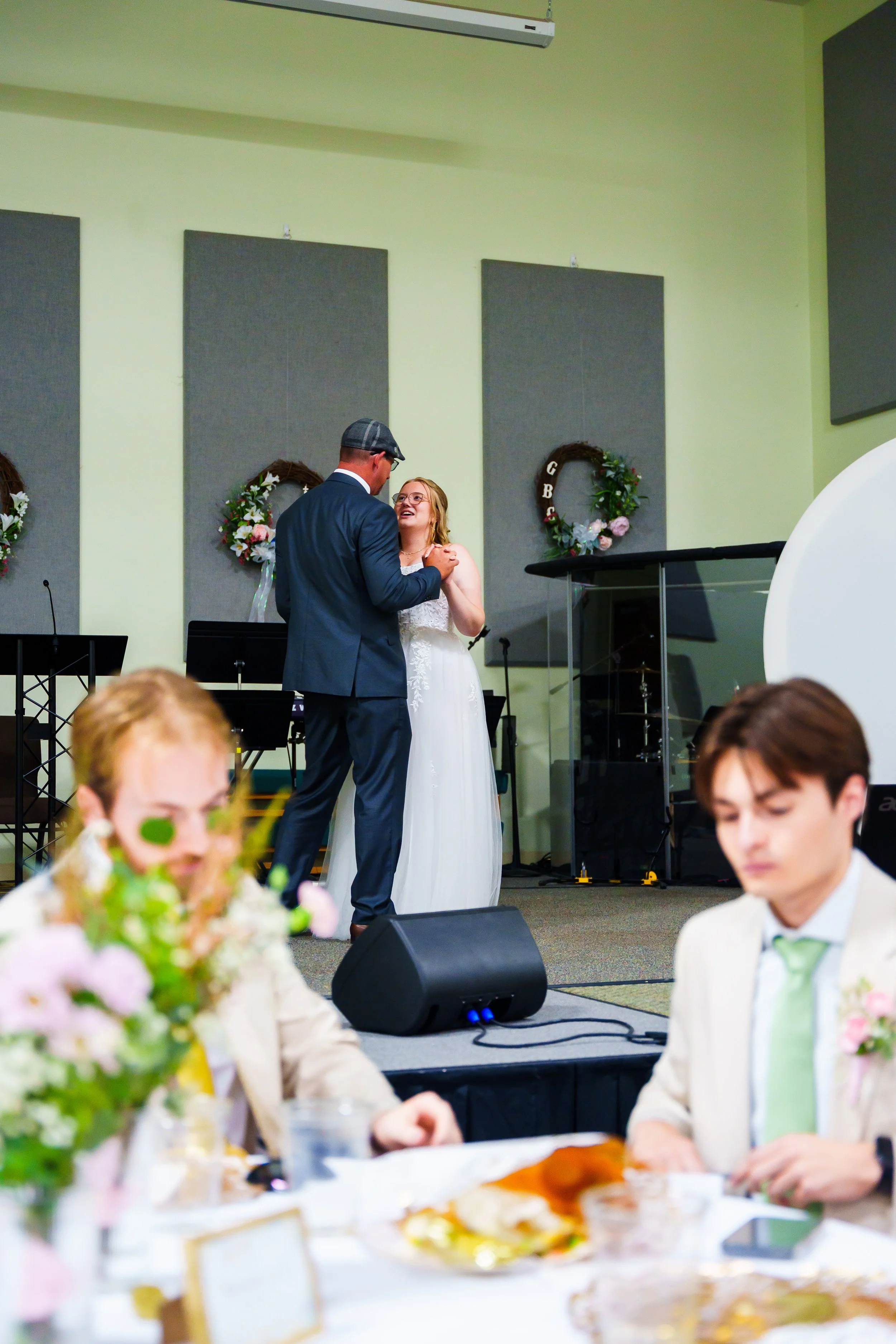 A bride in a white wedding dress and a groom in a blue suit dance closely together at a wedding reception in a decorated hall.