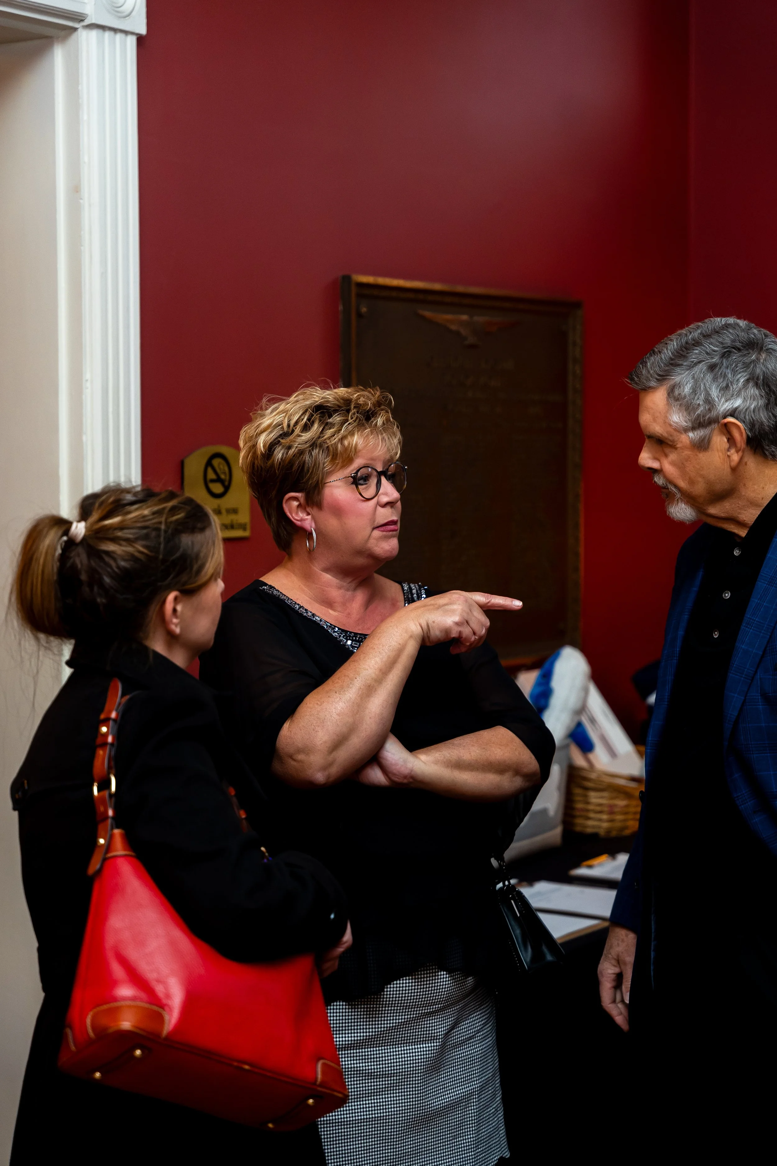 A woman with short curly hair and glasses is talking to a man with gray hair and a beard, pointing her finger. A young girl with a ponytail and a woman with a red bag are also in the scene. They are indoors with a red wall and a framed plaque in the 