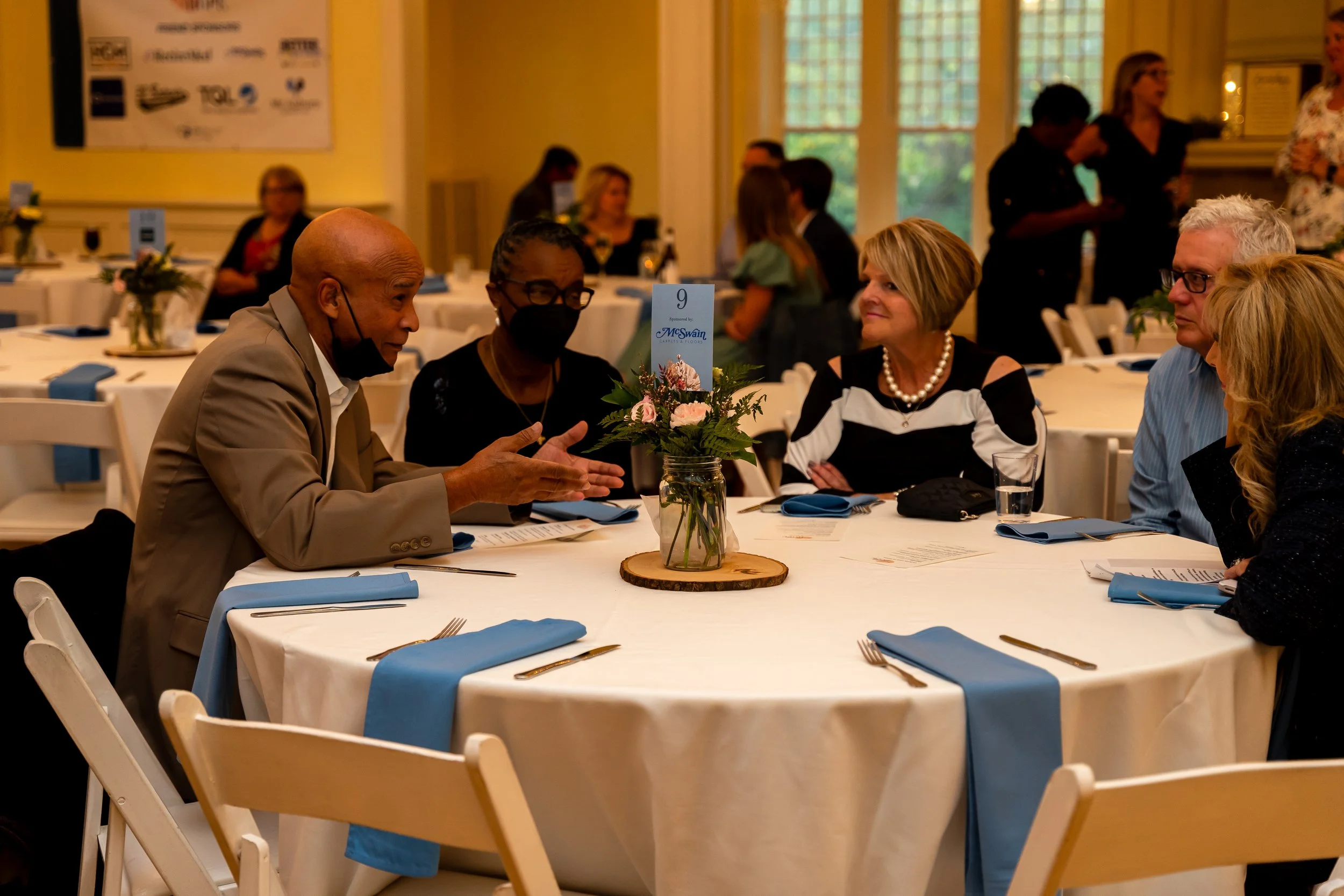 People sitting at round tables decorated with flowers, engaged in conversation at a formal event in a banquet hall with large windows.