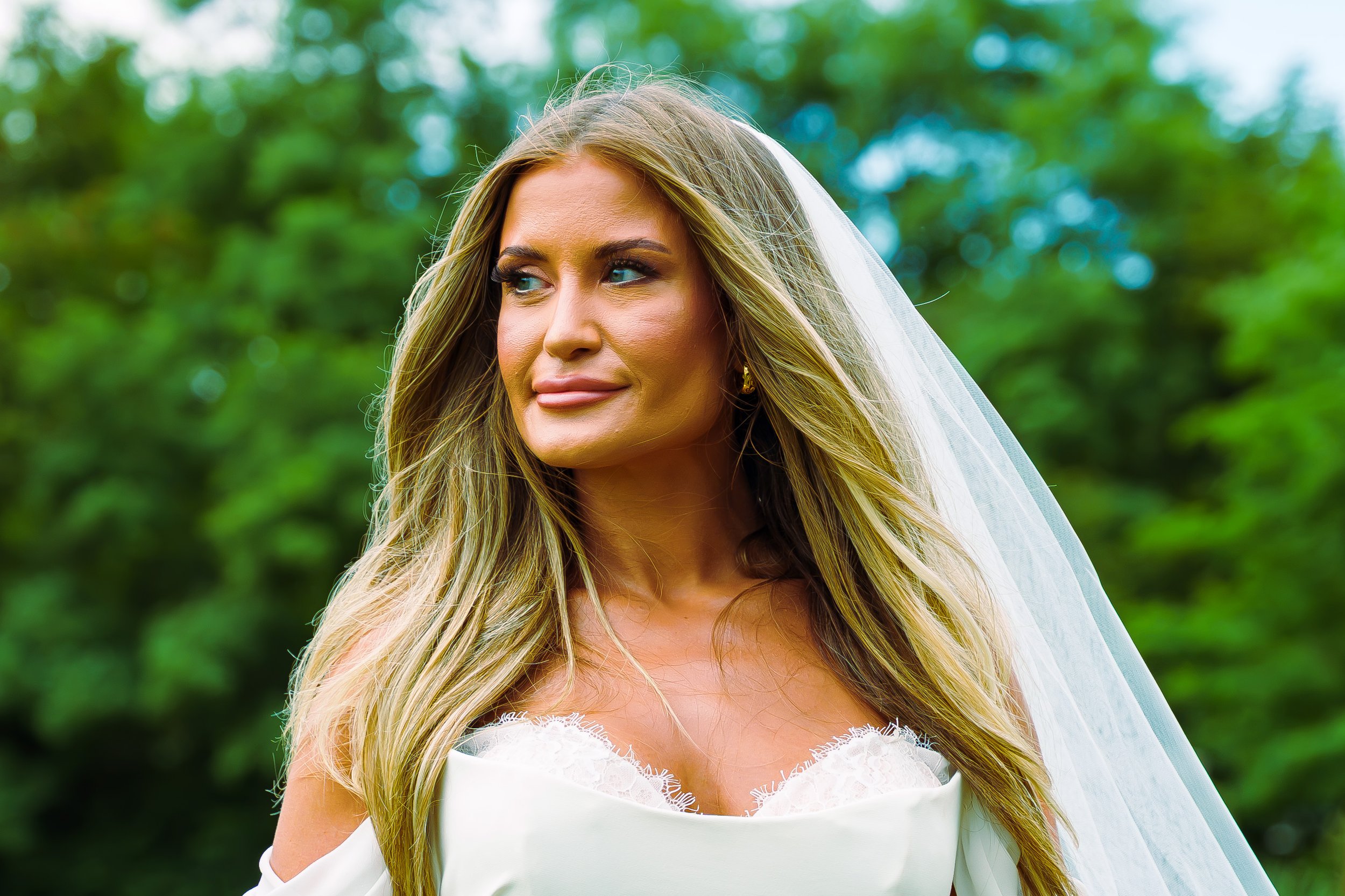 A woman with long blonde hair, wearing a white lace wedding dress and veil, standing outdoors with green blurred foliage in the background.