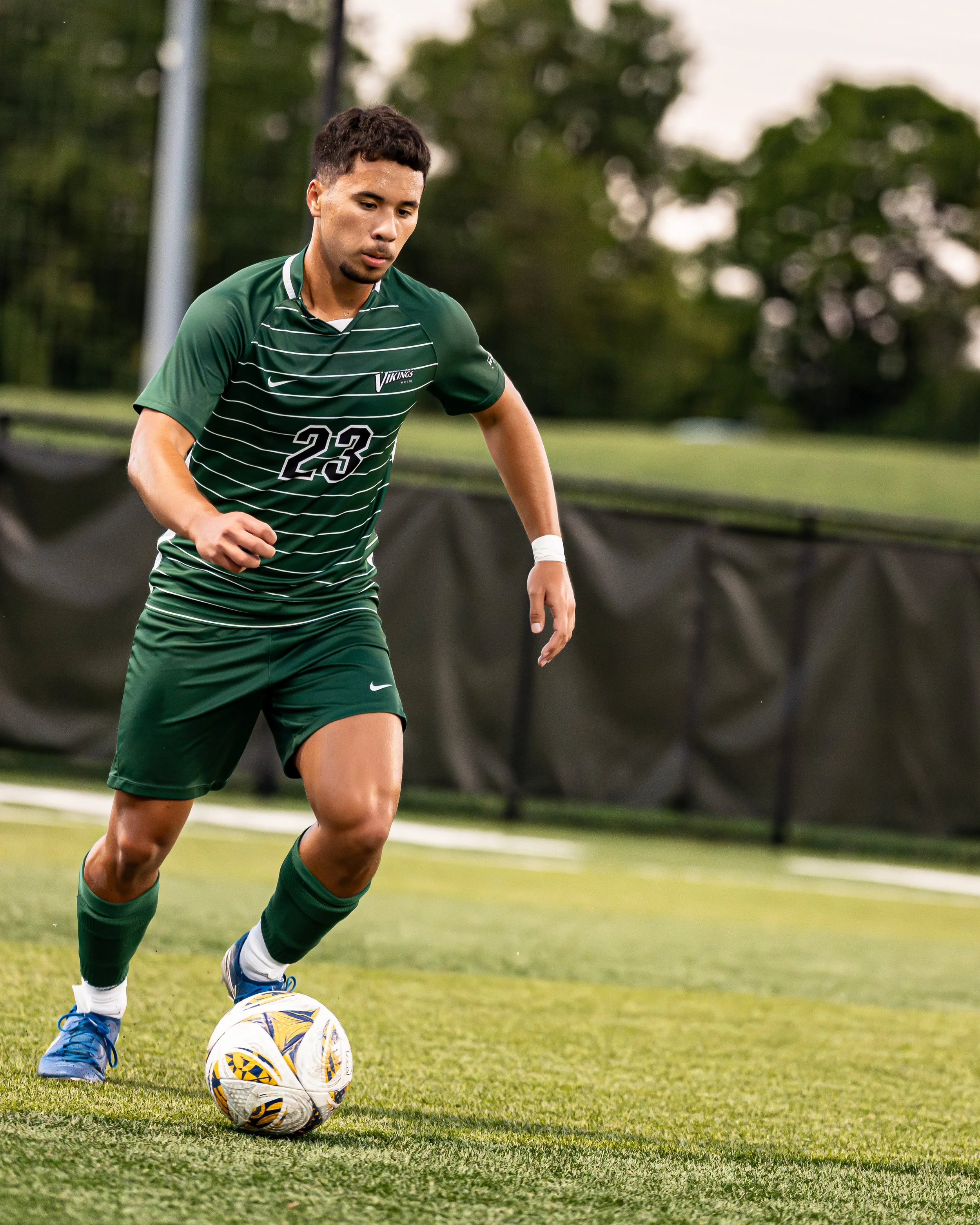 A male soccer player in a green uniform with the number 23 dribbling a soccer ball on a grassy field.