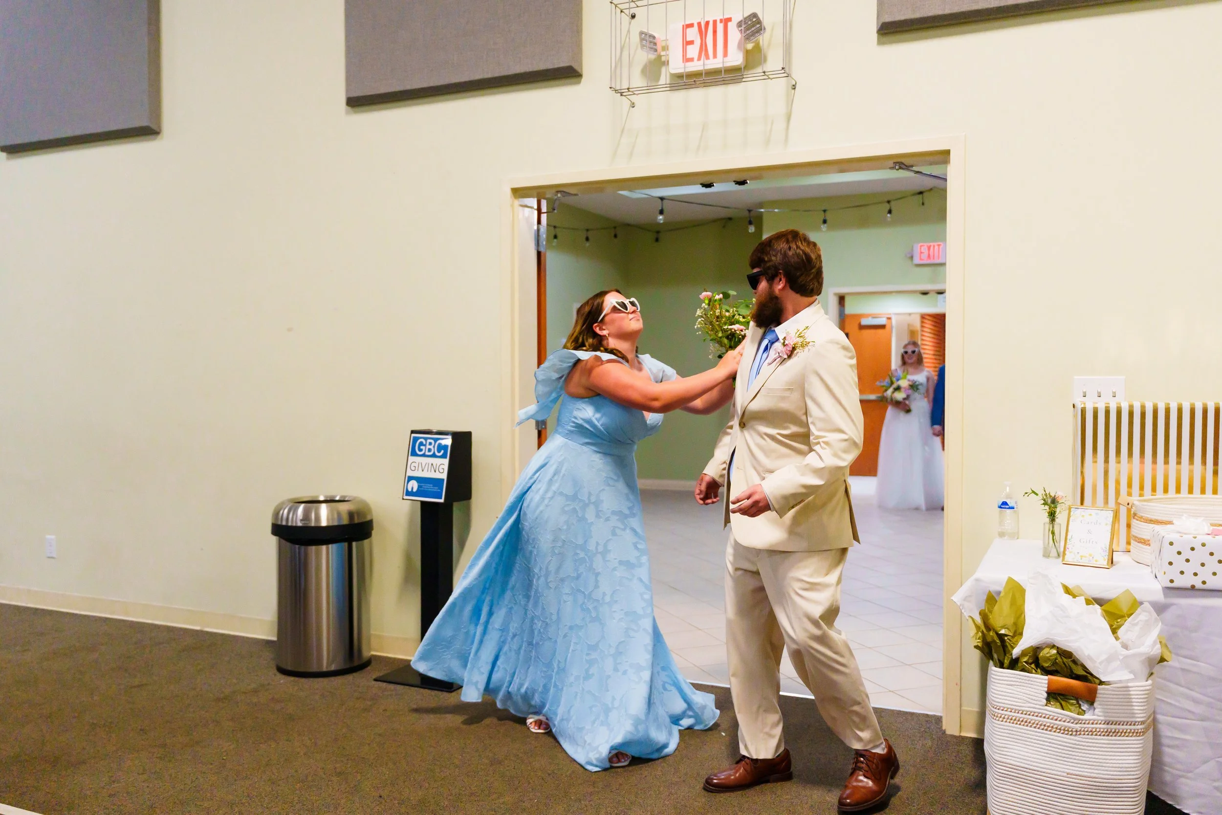 A woman in a light blue gown and sunglasses is touching the lapel of a man in a cream suit and sunglasses, with a woman in a white dress and sunglasses holding a bouquet in the background at a wedding reception.