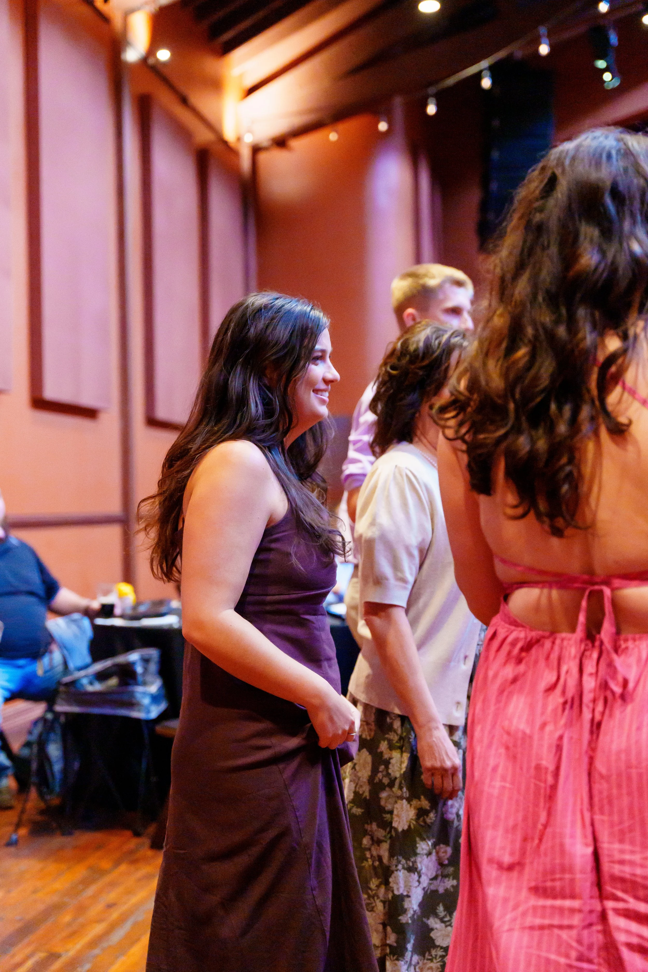 A group of women and men standing in a line indoors with wooden ceiling beams, engaged in a dance or activity, smiling and facing forward, with warm lighting and painted walls in the background.