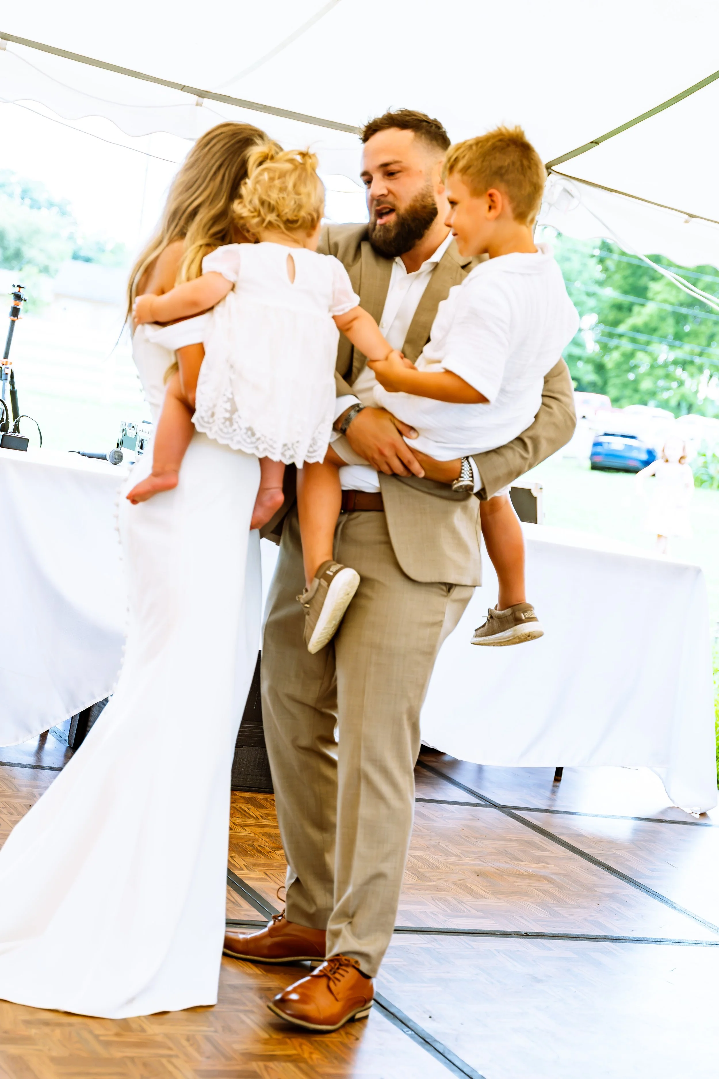 A man in a beige suit holding two young children, one girl and one boy, at an outdoor event under a white tent.