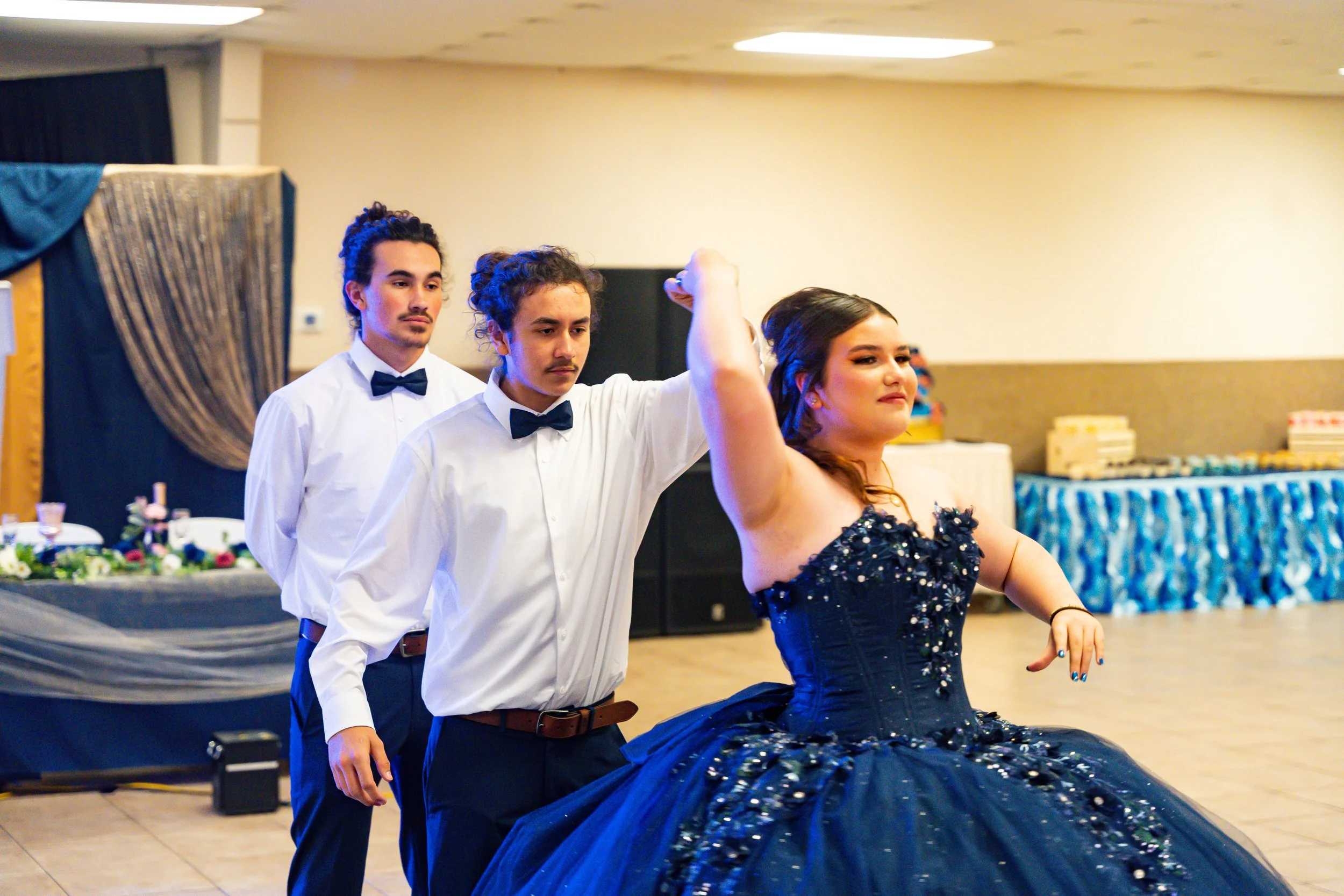 Three young adults in formal attire dancing at a celebration or event in a banquet hall. The men are wearing white shirts and bow ties, while the woman is in a dark blue dress. They are performing a dance with the woman seated and the men standing be
