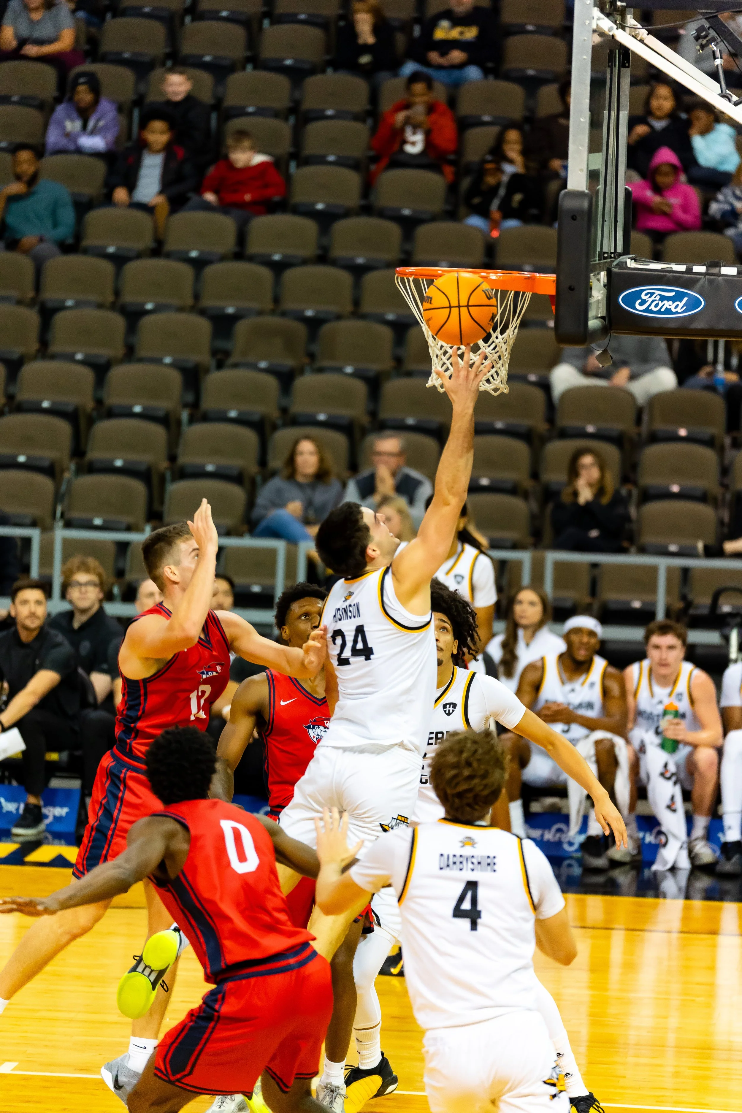 A basketball player in a white jersey leaps to score a basket in a game with opposing players in red jerseys attempting to block him.