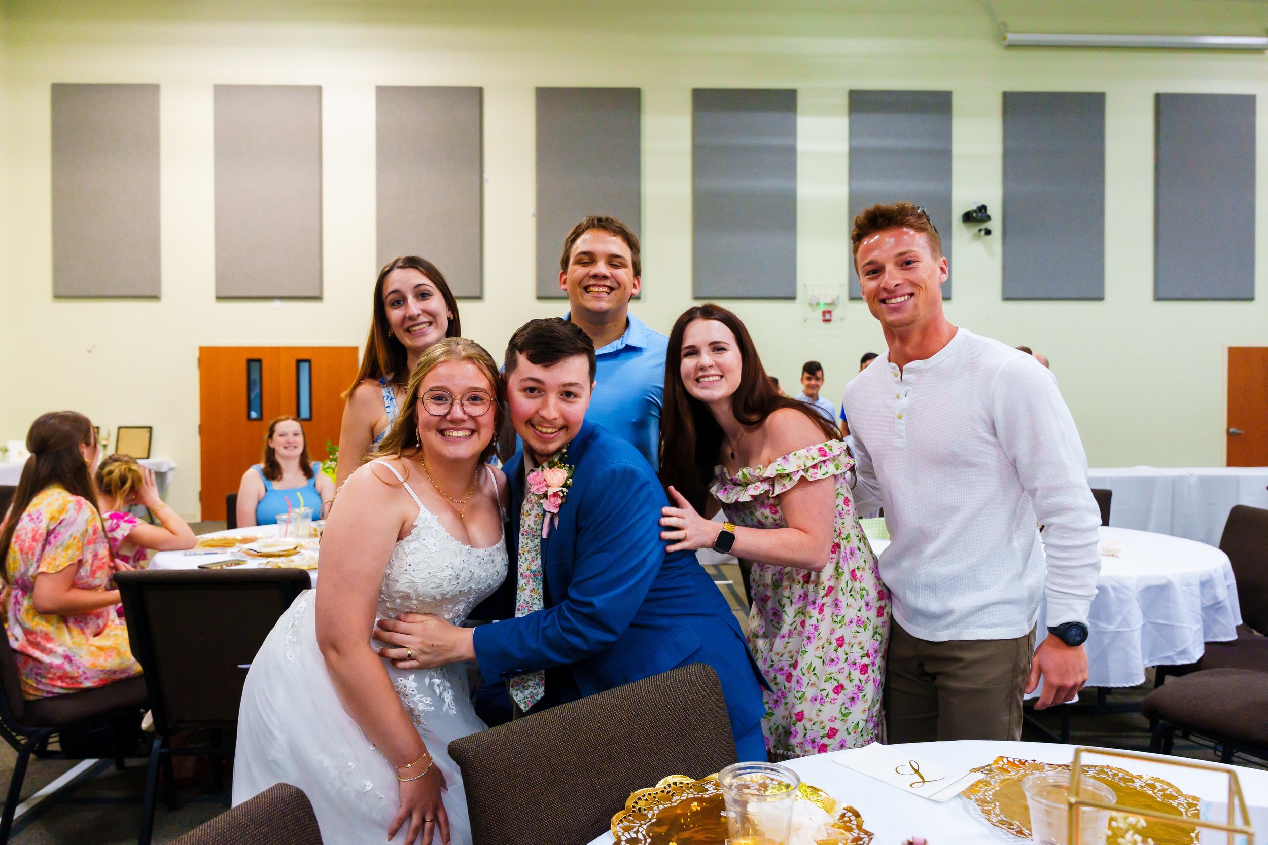 Group of six smiling young people, including a bride and groom, posing together at a celebration in a banquet hall.
