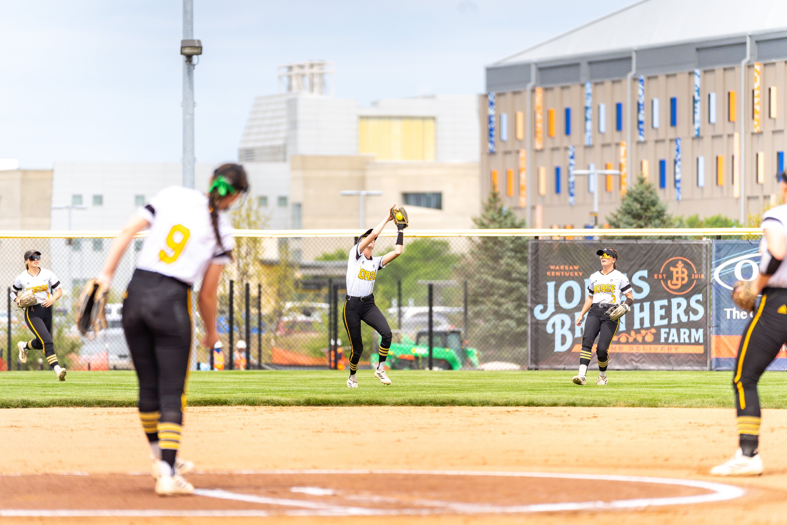 Soft-focus image of young women playing softball, with a field and colorful buildings in the background.