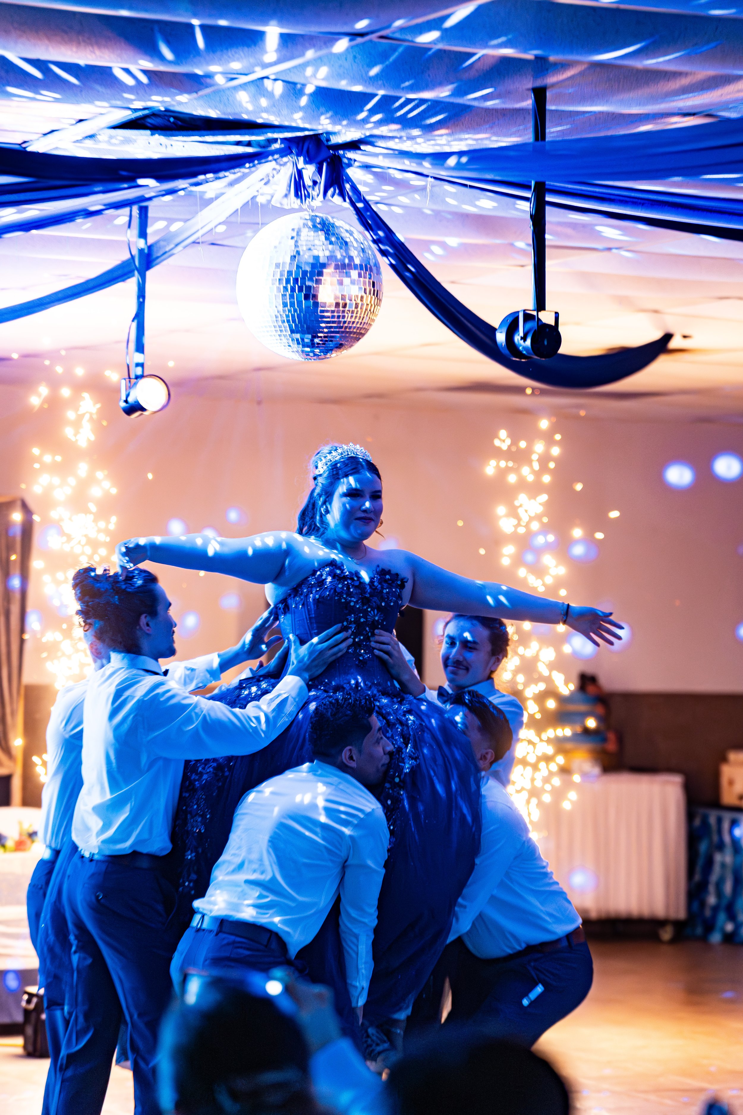 A group of people dancing at a celebration, with a woman being lifted by others, a disco ball hanging from the ceiling, and colorful lighting and sparklers in the background.