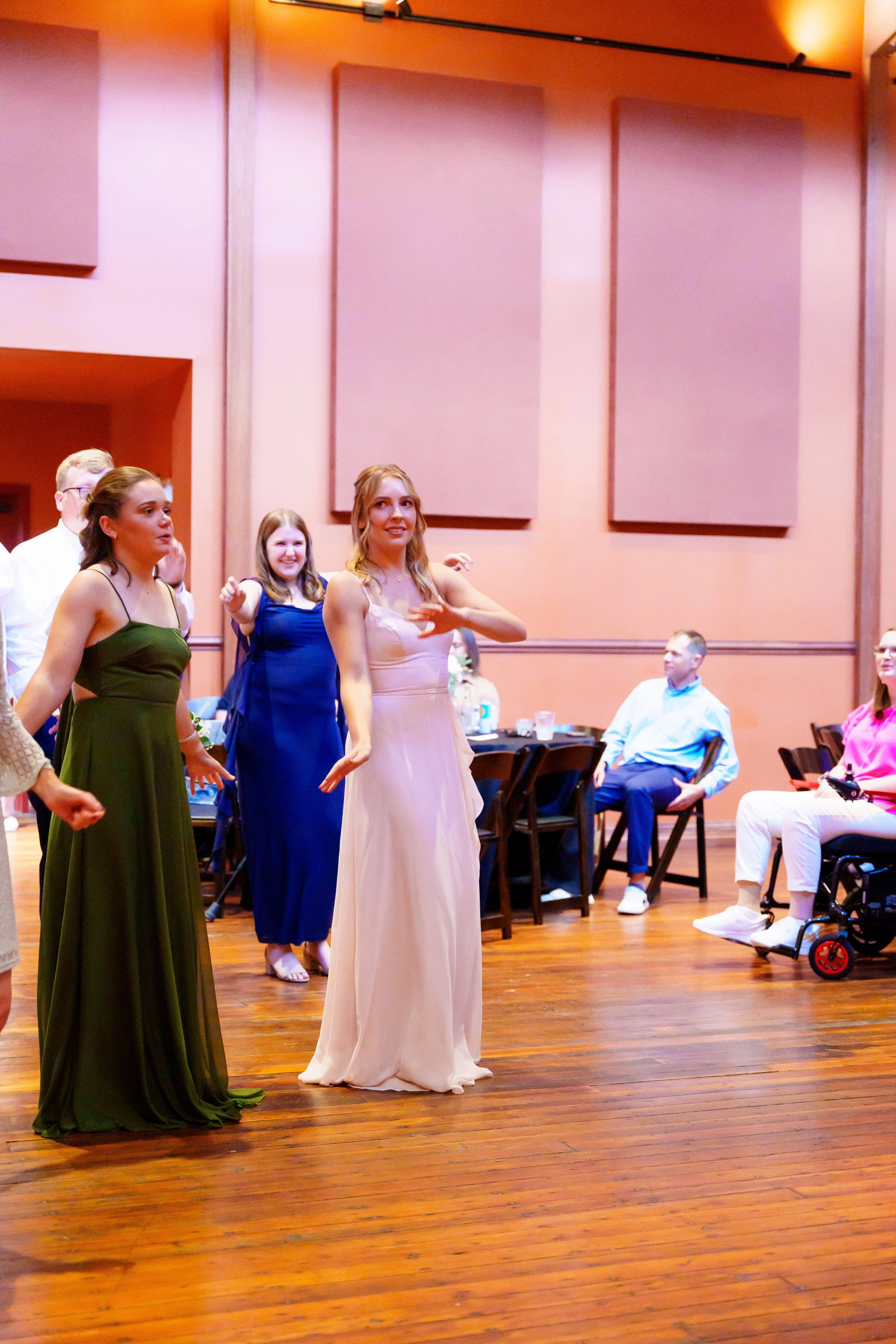 Group of young women in formal dresses standing on a wooden floor at an indoor event, some sitting at tables in the background.