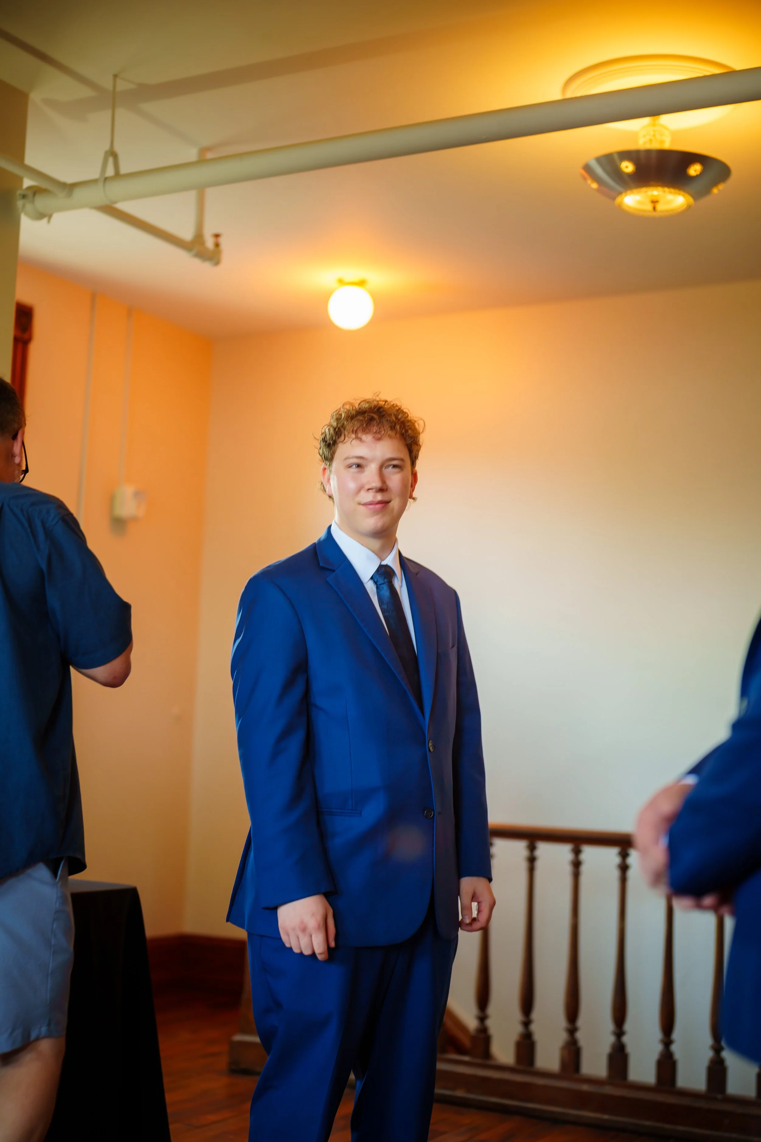 Young man in a blue suit looking to the side at an indoor event with warm lighting, a preserved wood staircase, and people around.
