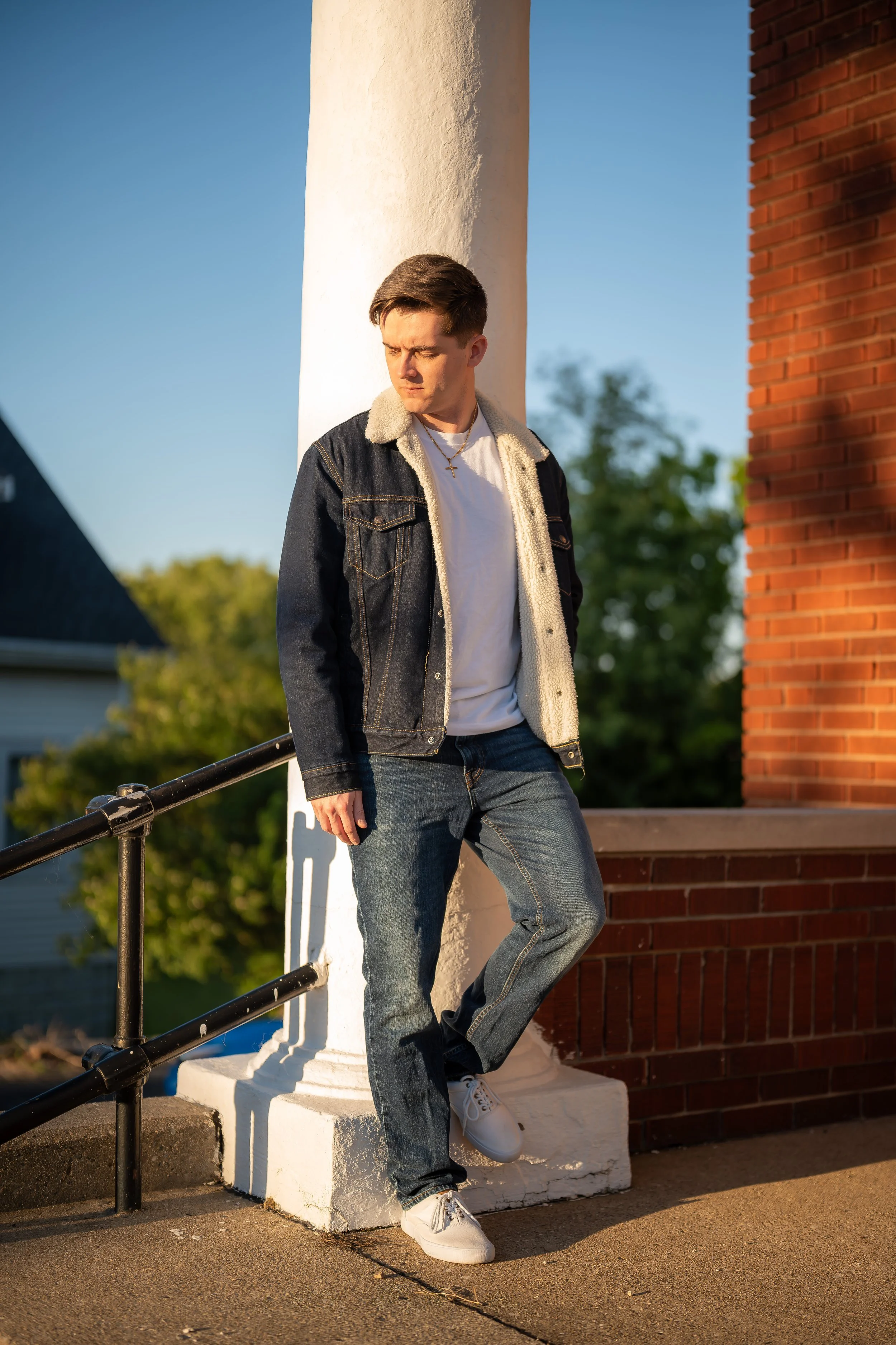 A young man with short brown hair wearing a denim jacket with a shearling collar, white t-shirt, and jeans standing outdoors next to a white column and a brick wall during sunset, with a serious or contemplative expression.
