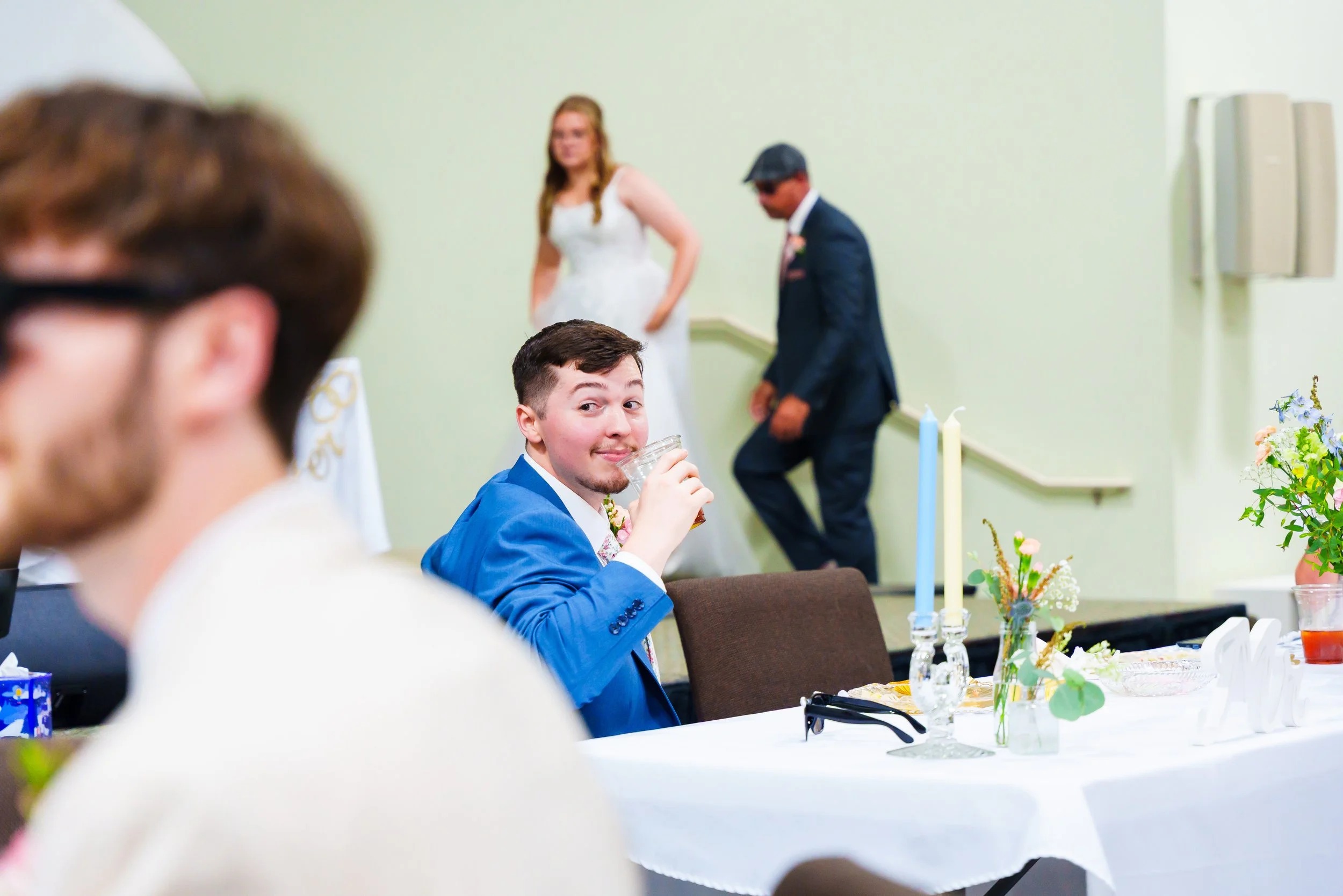 A young man in a blue suit sitting at a decorated table during a celebration, smiling and holding a glass, in a room with light green walls, flowers, and several candles.