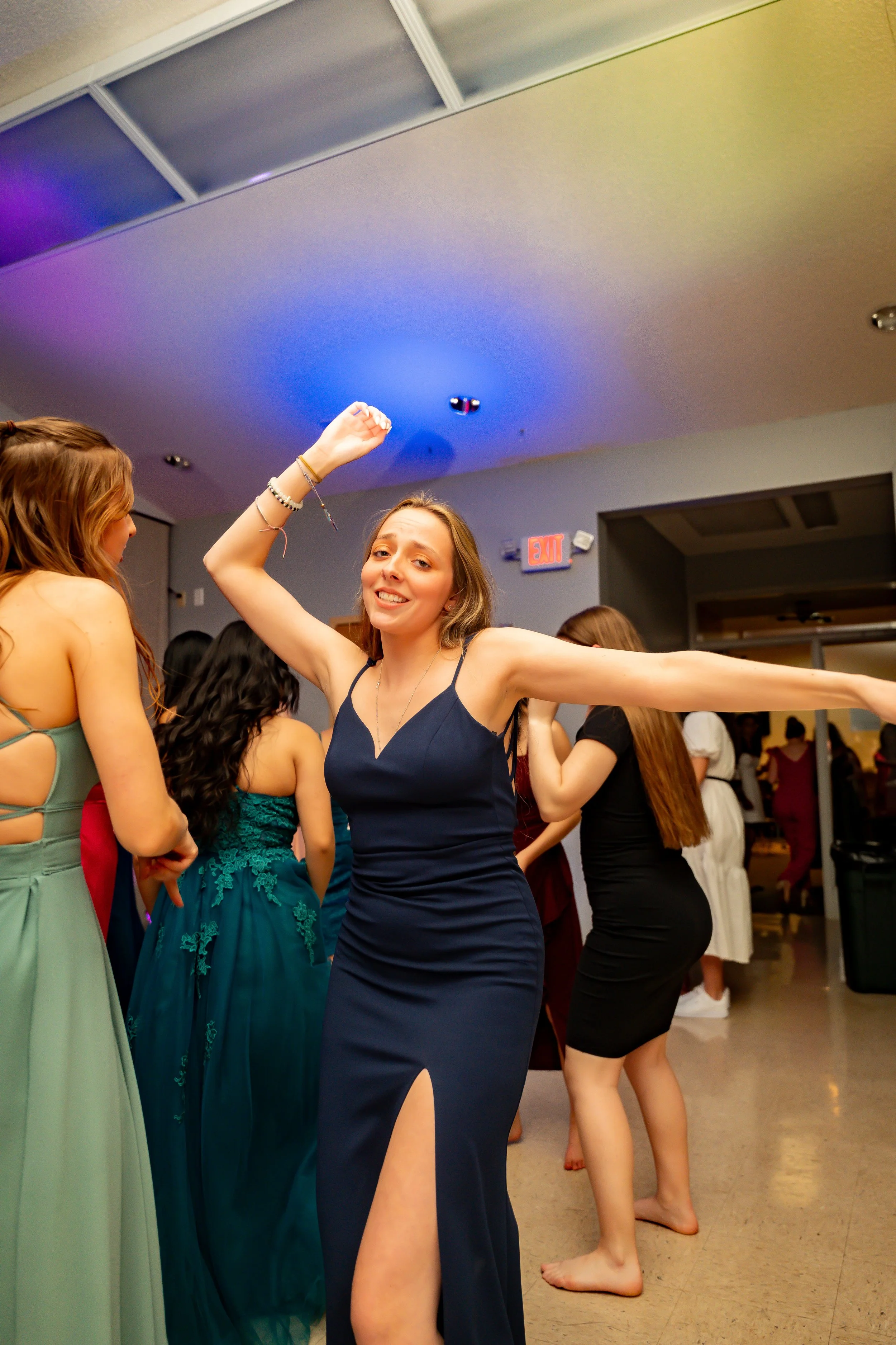 A group of women wearing formal dresses dancing at an indoor event. The woman in the center is smiling, wearing a navy blue dress with a thigh-high slit, and raising her arm.