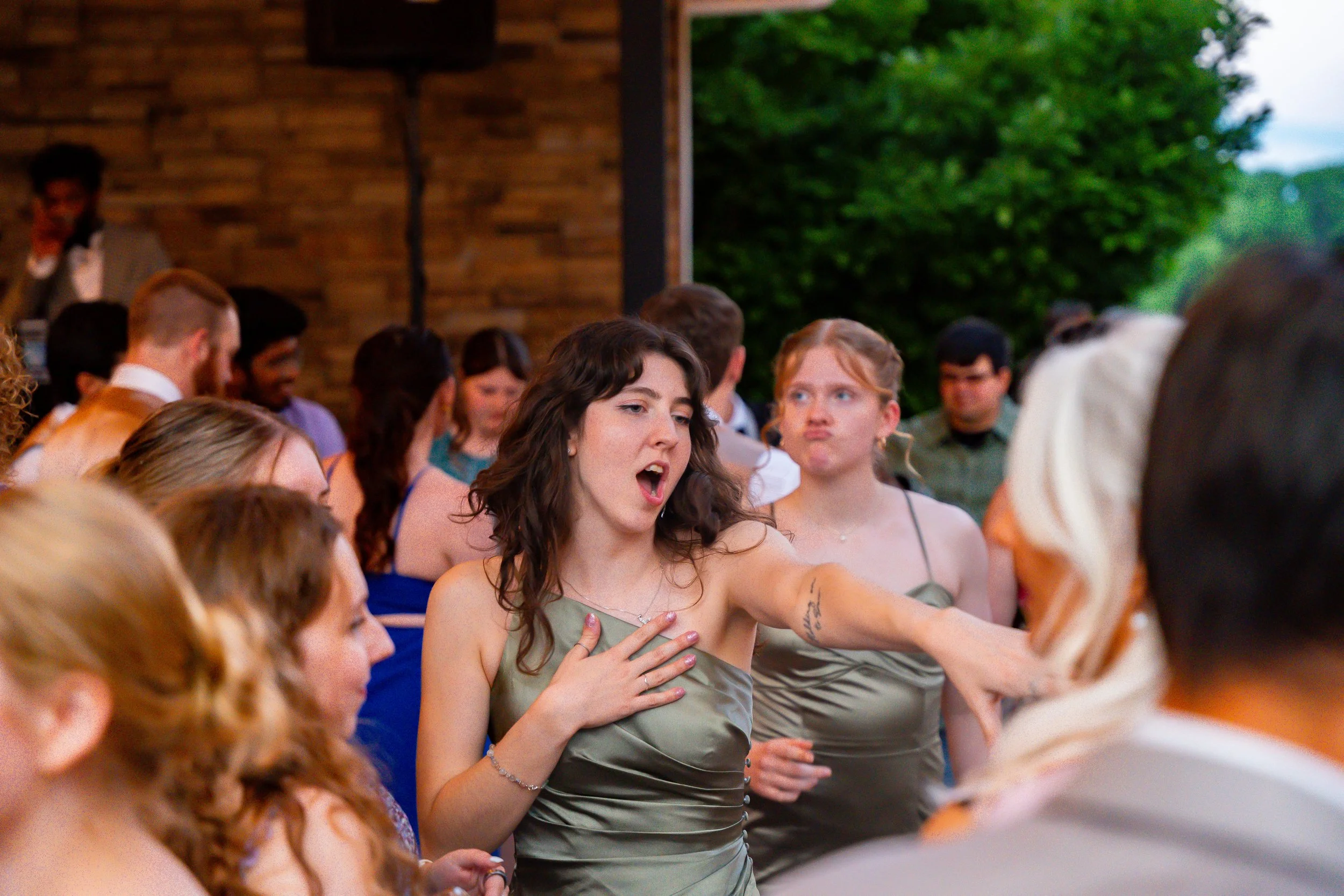 A group of people at an outdoor event, with a woman in the center touching her chest and speaking, wearing a gray dress. The background has a wooden wall and green trees.
