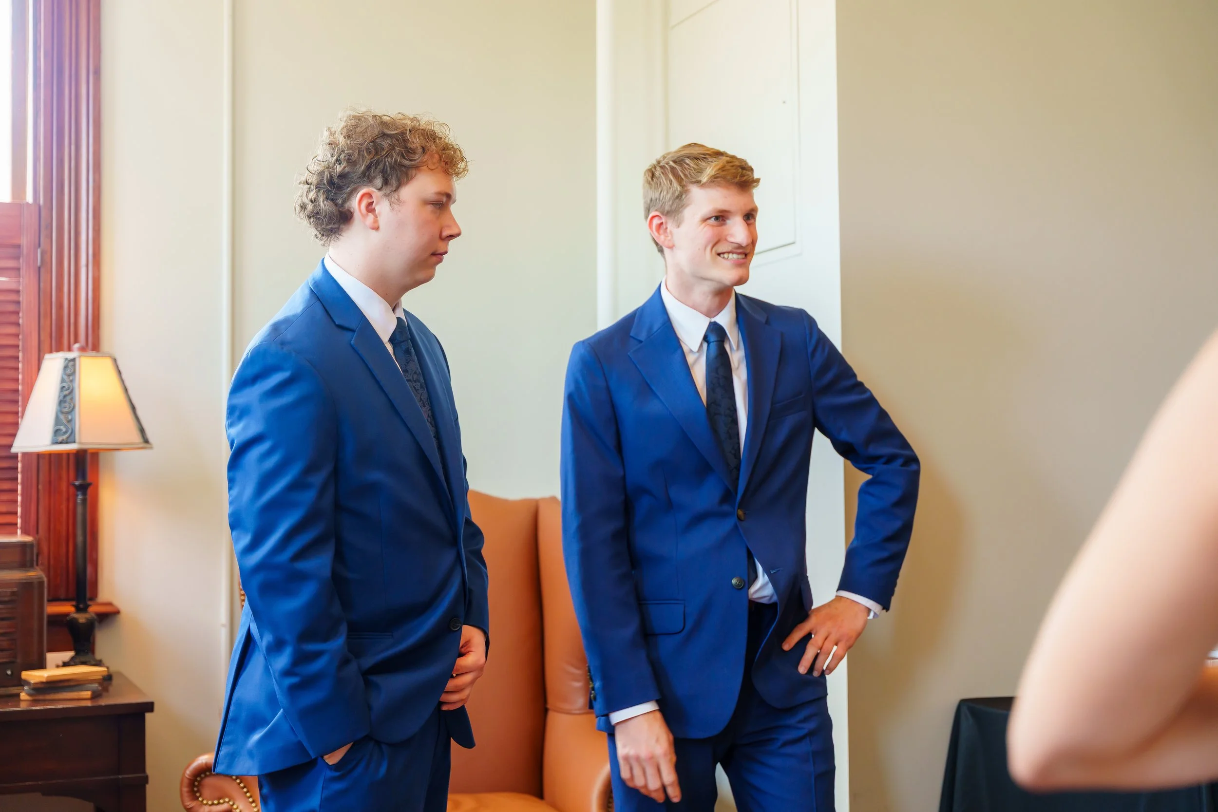 Two young men in blue suits standing indoors, engaged in conversation, with neutral-colored walls, a wooden side table with books, and a table lamp in the background.