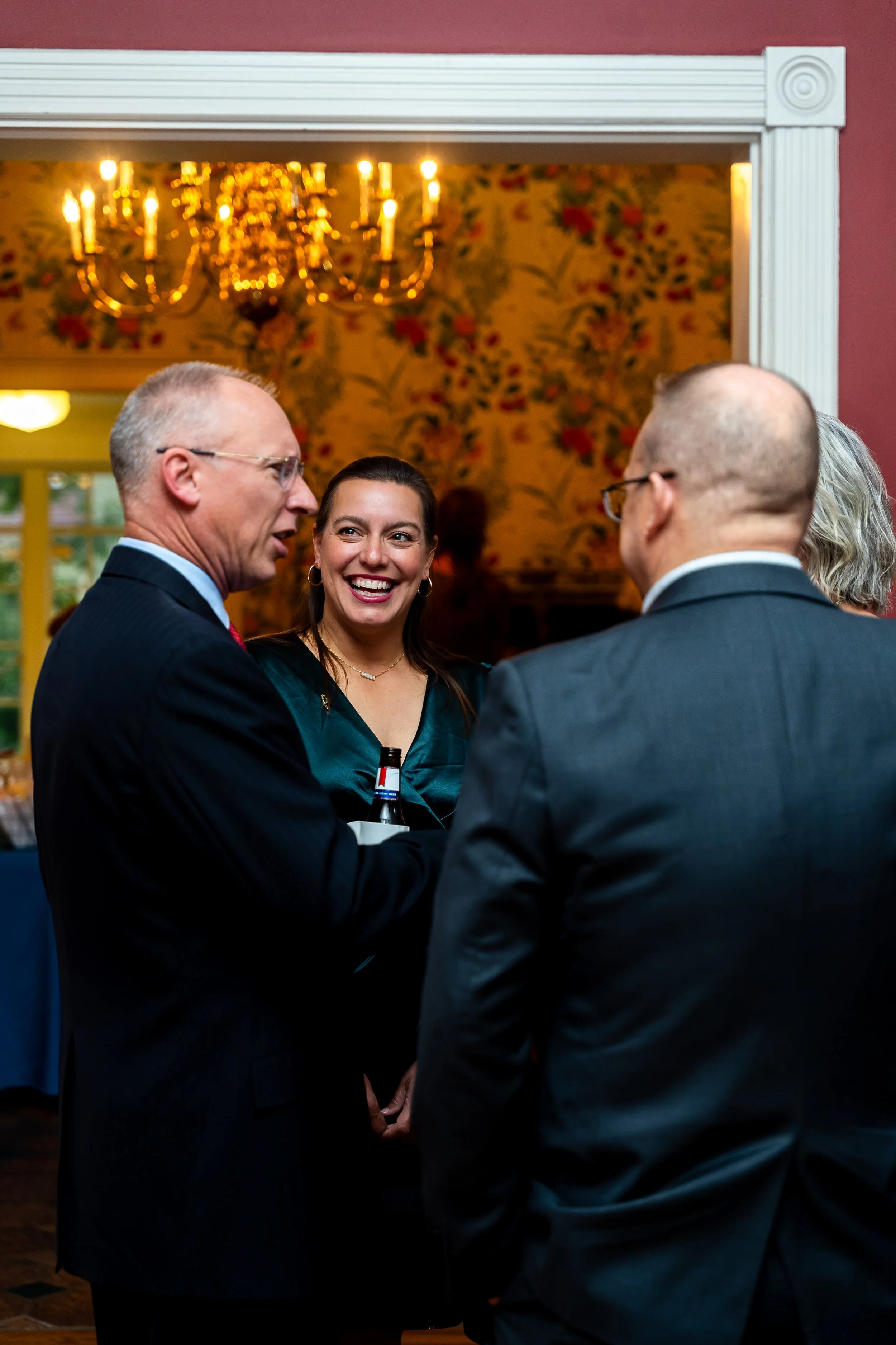 People in formal attire engaged in conversation at a social event, with a chandelier and floral wallpaper in the background.