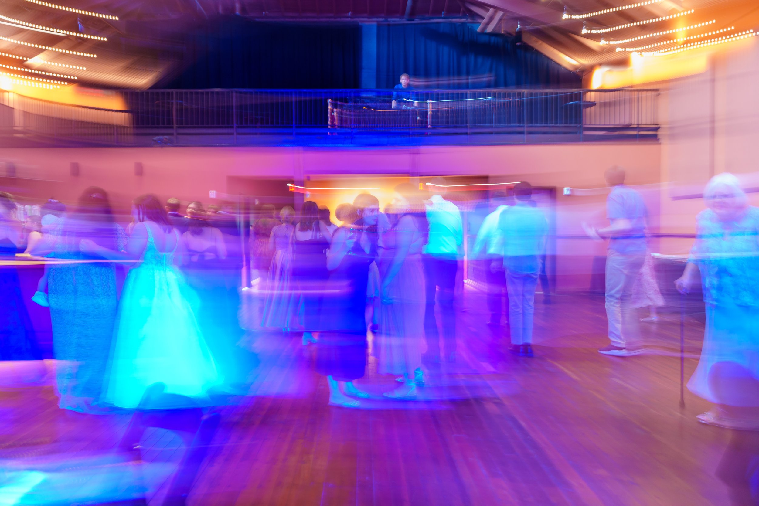 A dance floor with people dancing, illuminated by colorful lights with a DJ standing at DJ equipment in the background.
