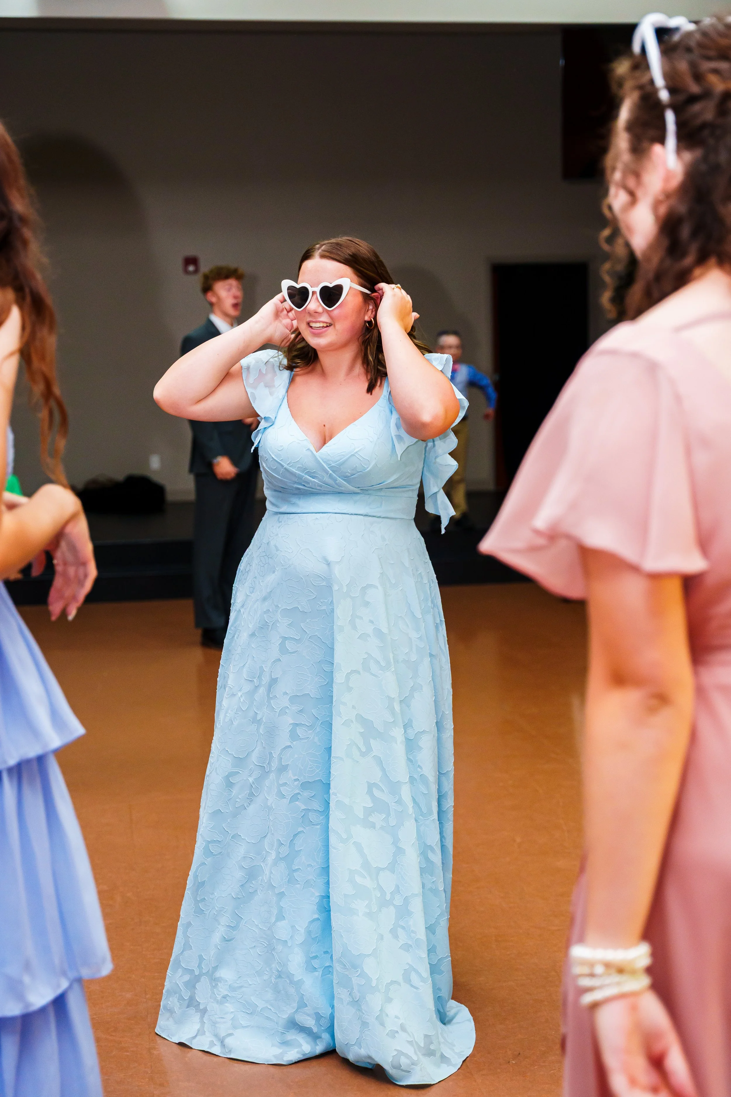 Woman in a light blue wedding dress adjusting heart-shaped sunglasses while standing on a dance floor at a celebration.