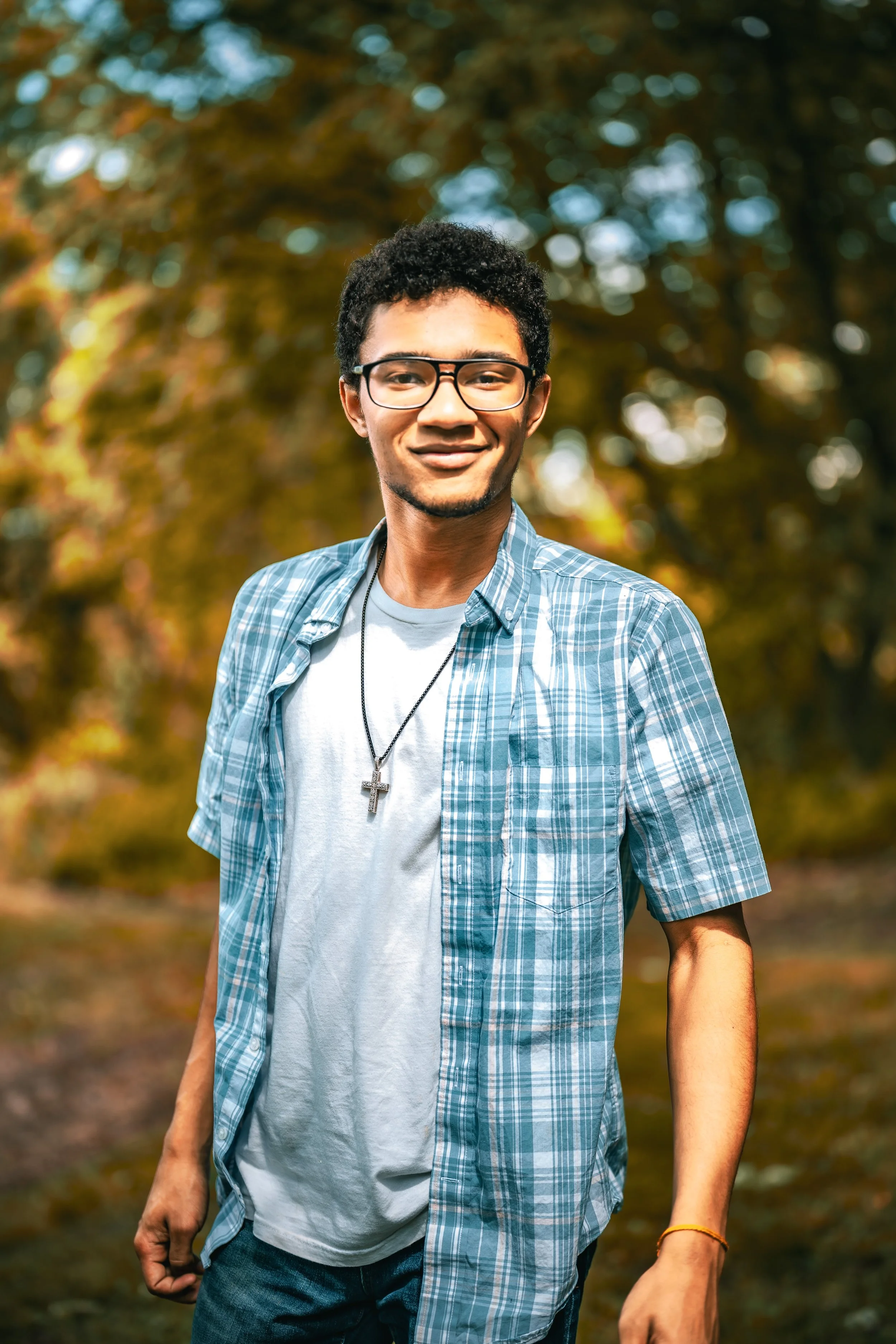 A young man wearing glasses, a blue plaid shirt over a white t-shirt, and a cross necklace walking outside in a park with autumn trees in the background.