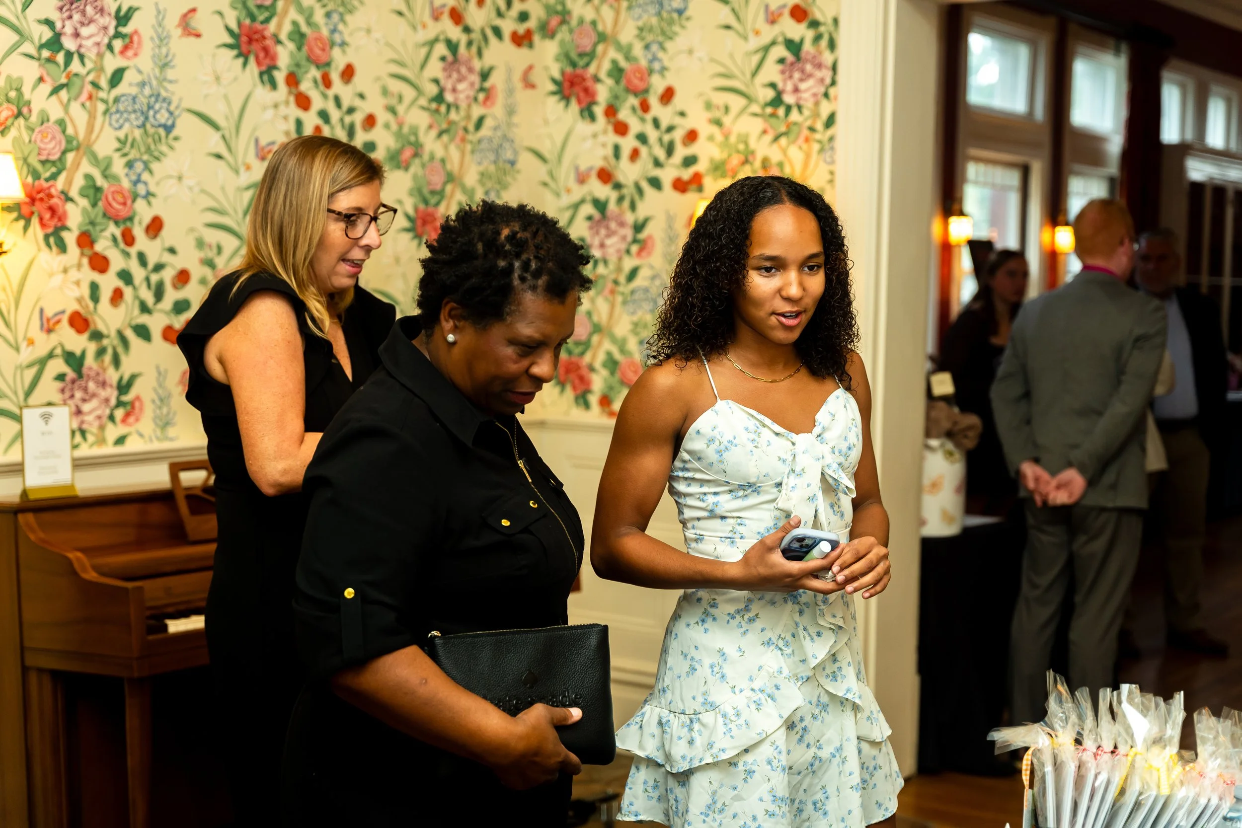 Three women standing together in a room with floral wallpaper, engaged in conversation. One woman with glasses and a black dress looks down, another woman with short curly hair and earrings looks down, and a young woman in a white dress with blue flo