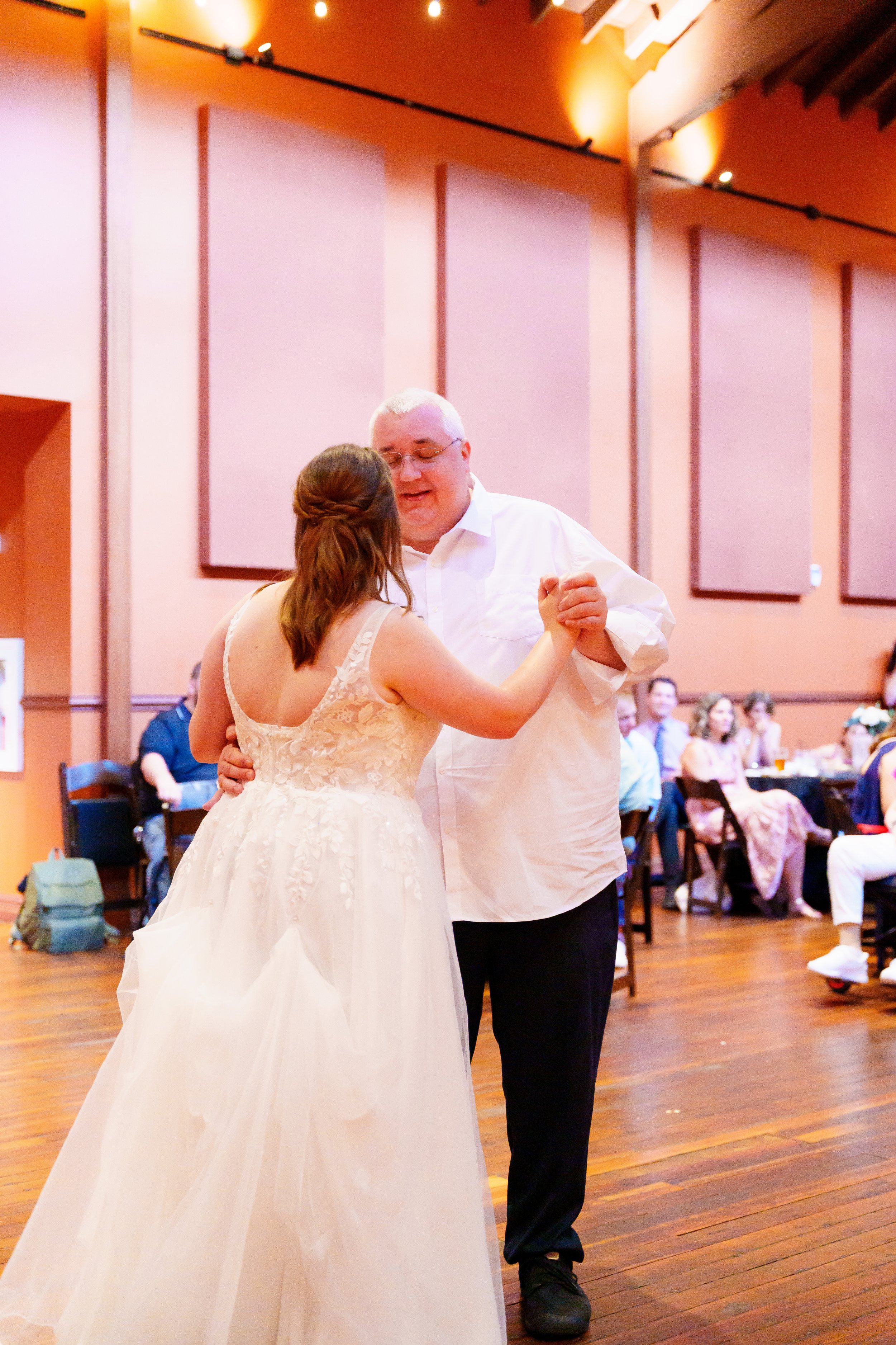 A couple dancing at a wedding reception in a decorated banquet hall with seated guests in the background.