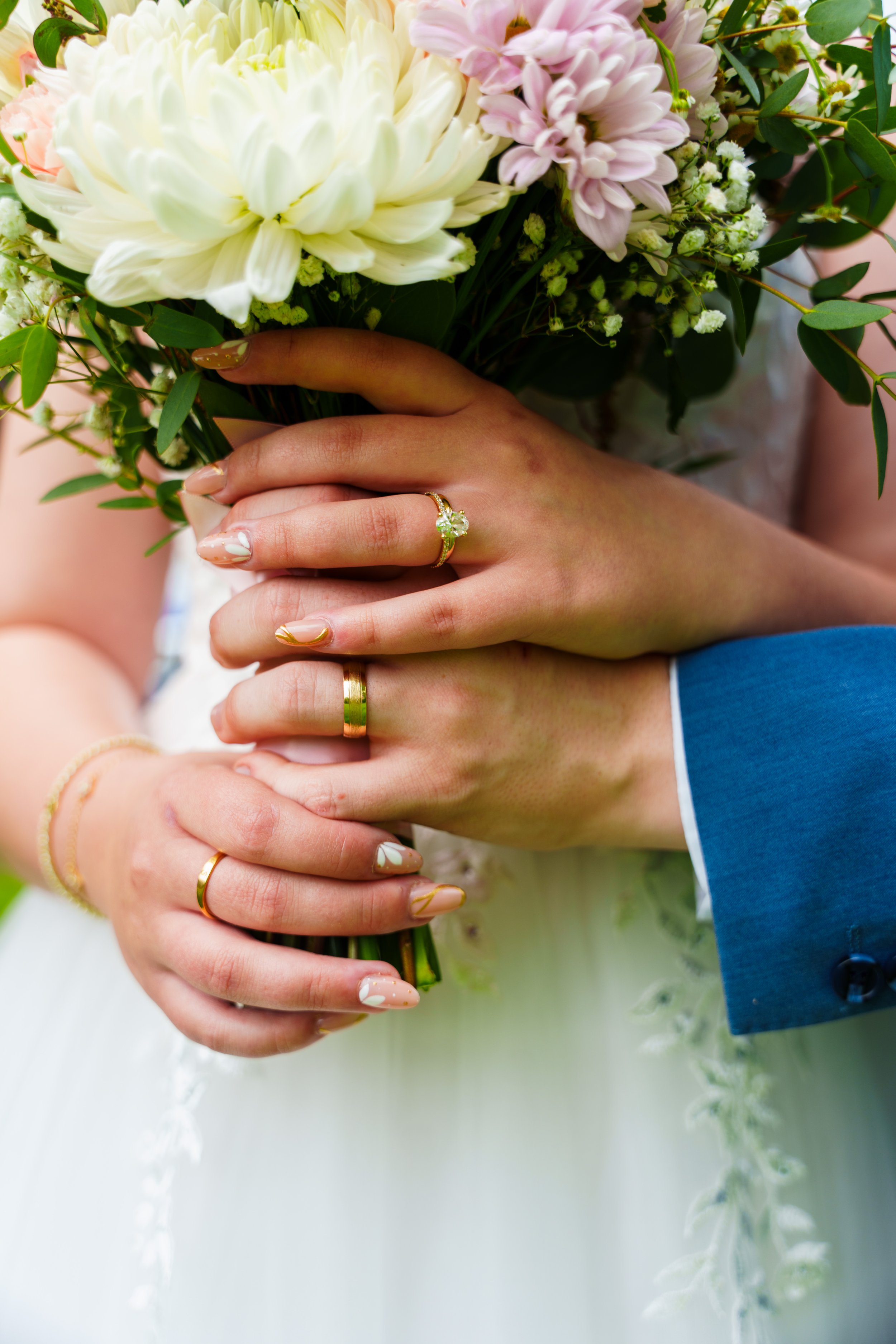 Close-up of a bride and groom's hands holding a bouquet of white and pink flowers, with bridal and groom rings visible.