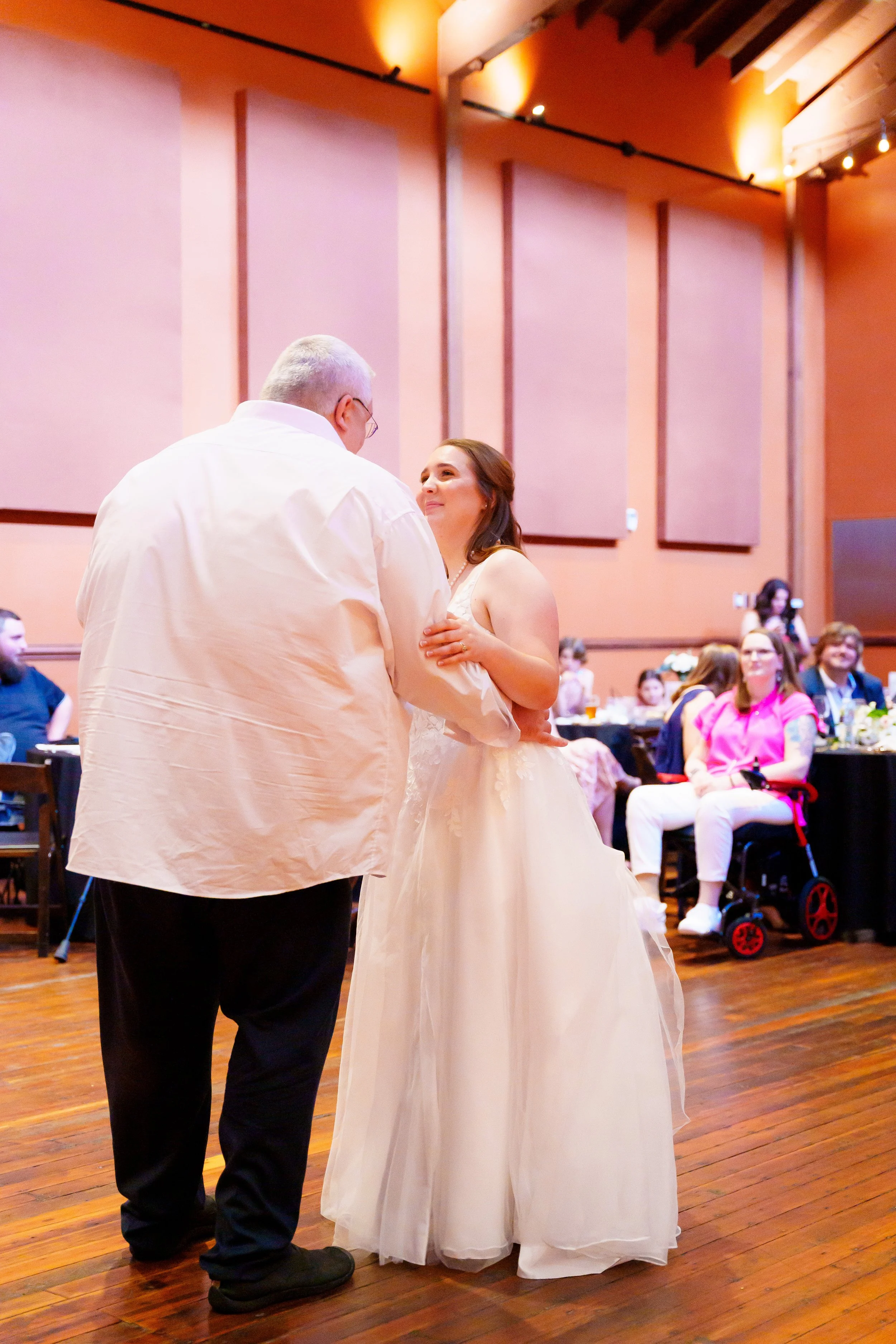 A couple dances at their wedding reception in a large hall with wood flooring and pink walls, with guests seated at tables in the background.