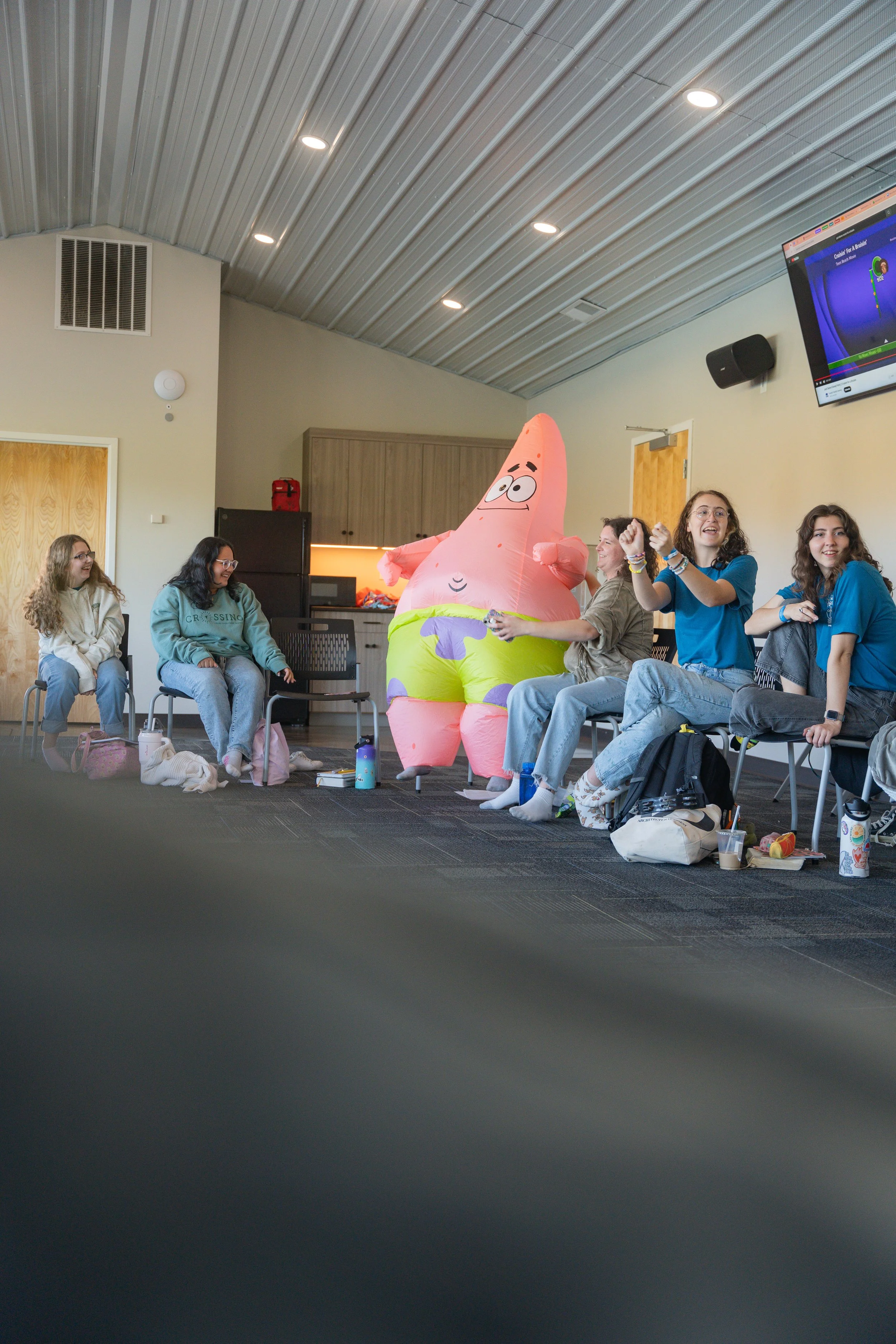 A group of young women sitting in a circle in a room with a ceiling of metal panels. One woman is dressed in a colorful inflatable Patrick Star costume from SpongeBob SquarePants. Some of the women are laughing and smiling, and there are various pers