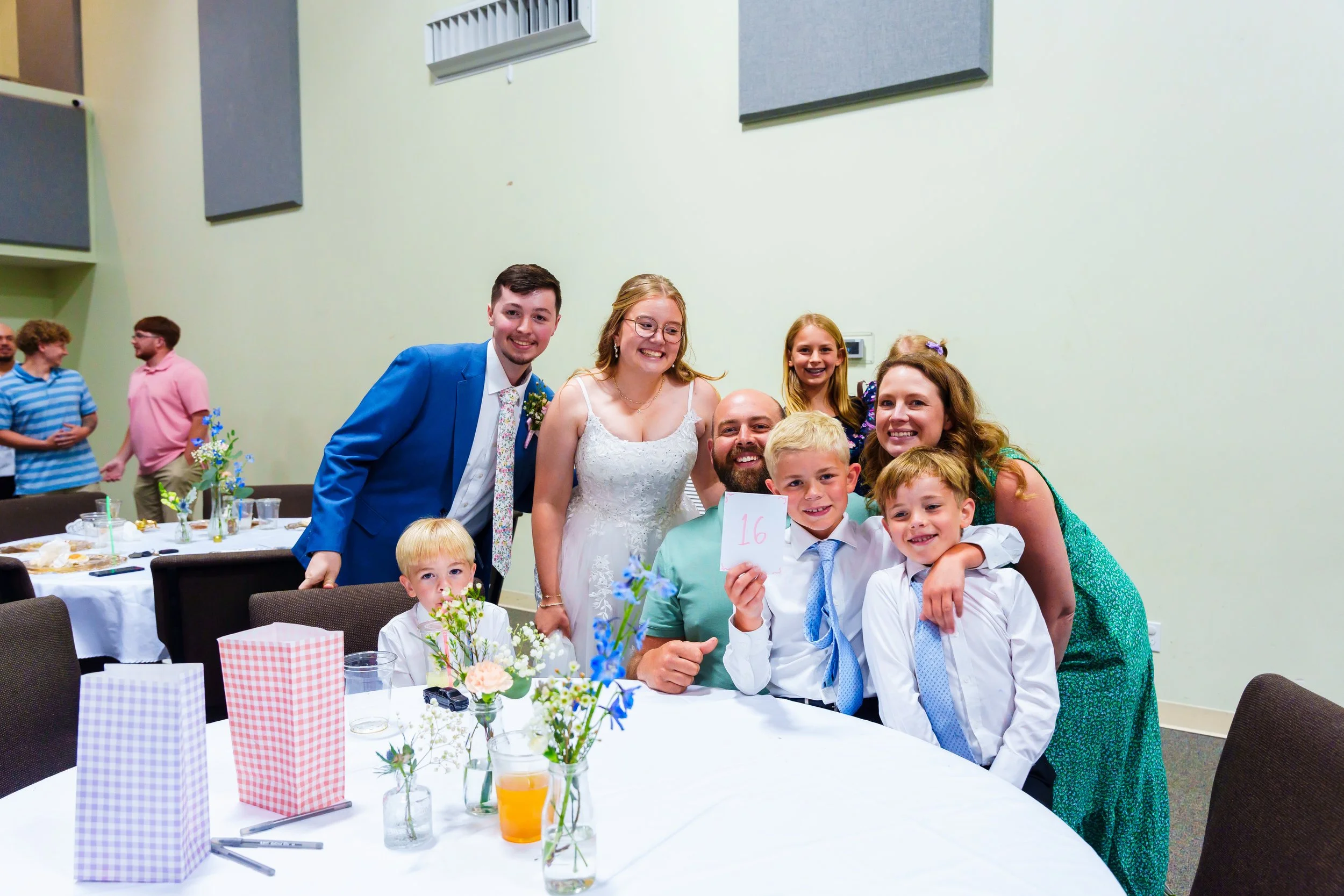 Group of people gathered around a table at a celebration or party, smiling and posing for a photo, with colorful flowers and table settings in the foreground.
