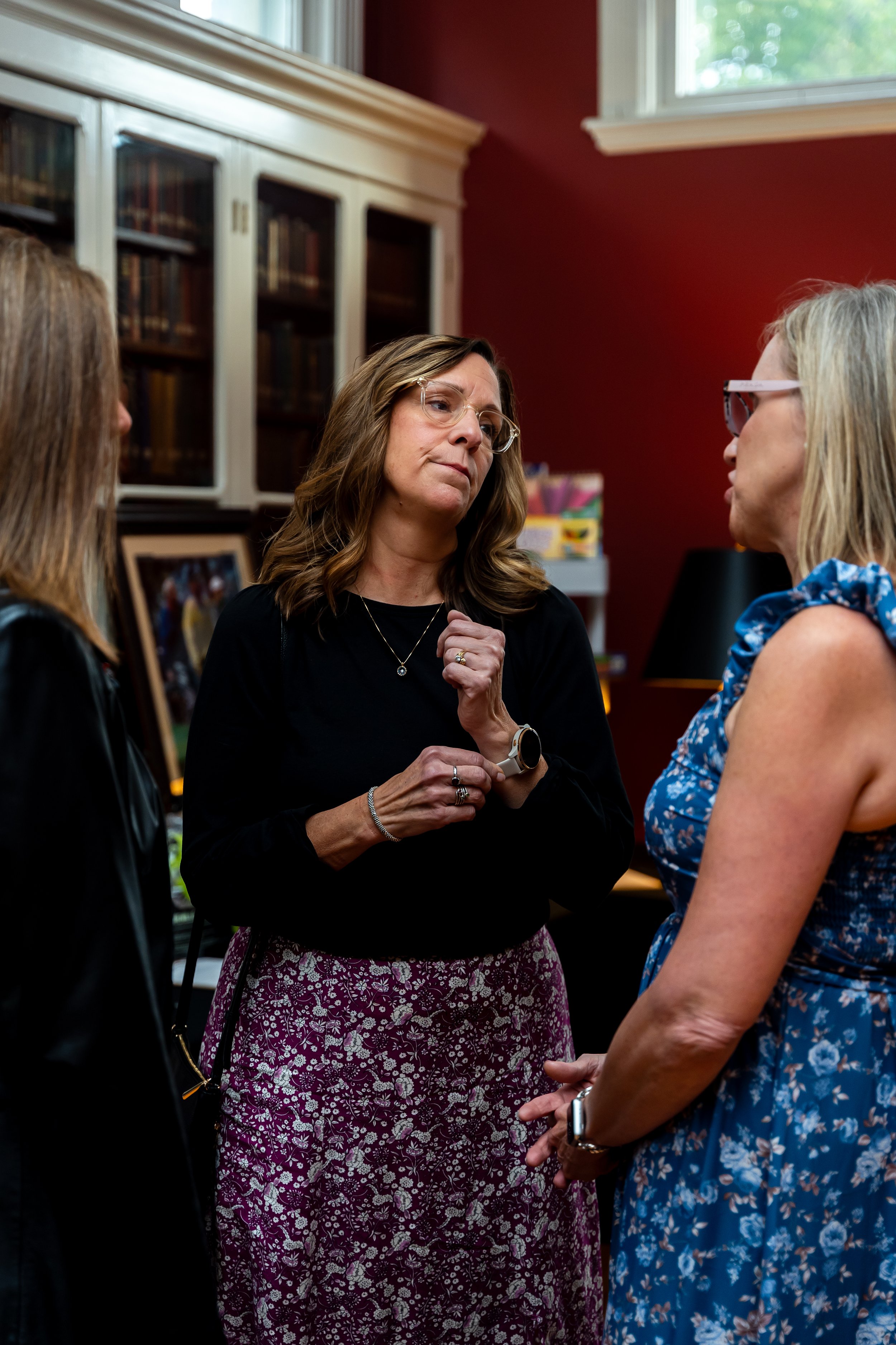 Three women in conversation in a room with a red wall, bookcase, and window. The woman in the center has wavy brown hair, wearing glasses, black top, patterned skirt, and gold jewelry. The woman on the right has blonde hair, glasses, and a blue flora