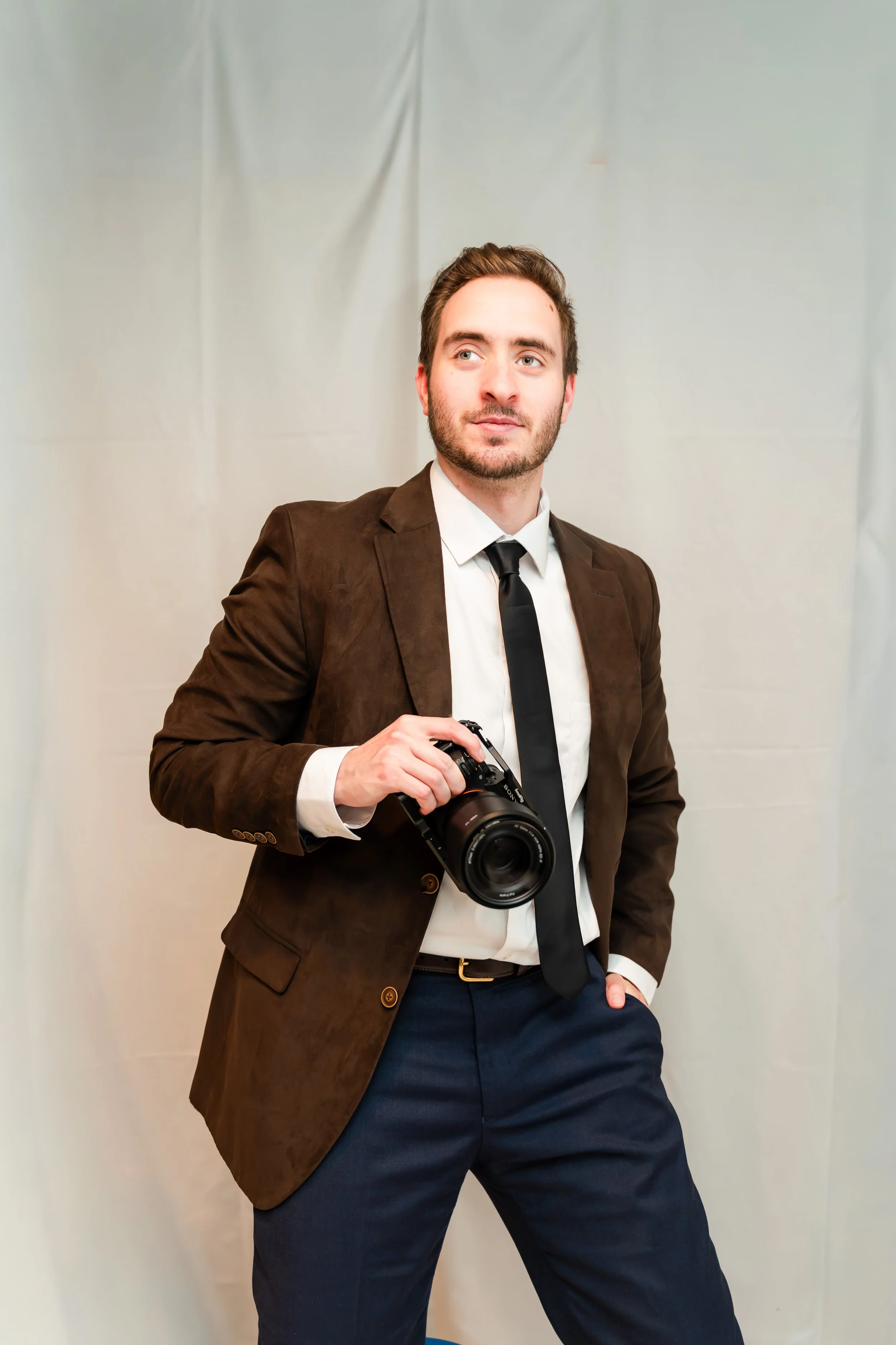 A young man in a brown suit jacket, white shirt, and black tie holding a camera stands against a plain light-colored backdrop.