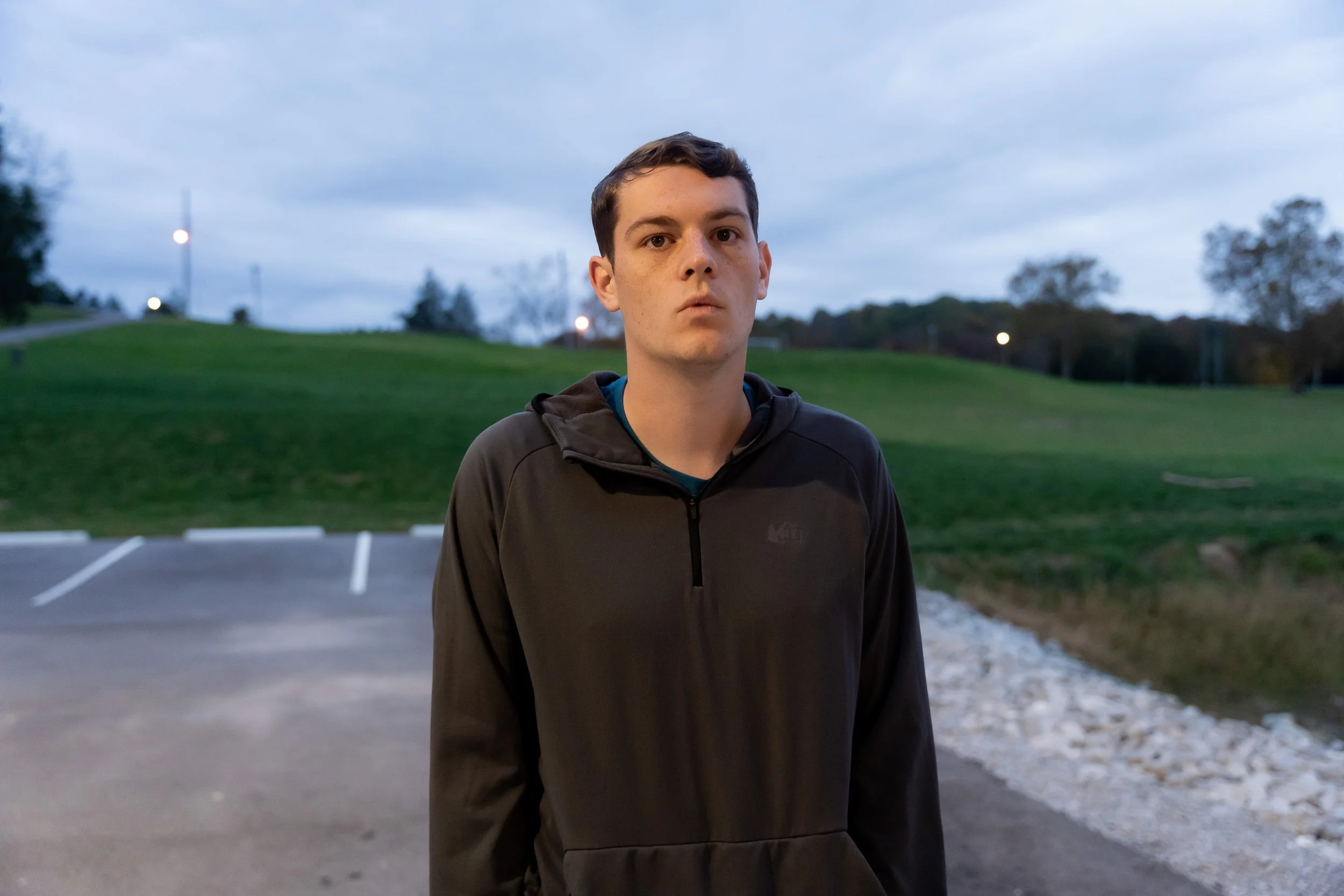 A young man with short dark hair and wearing a black jacket standing outdoors in a parking lot with trees and a cloudy sky in the background.